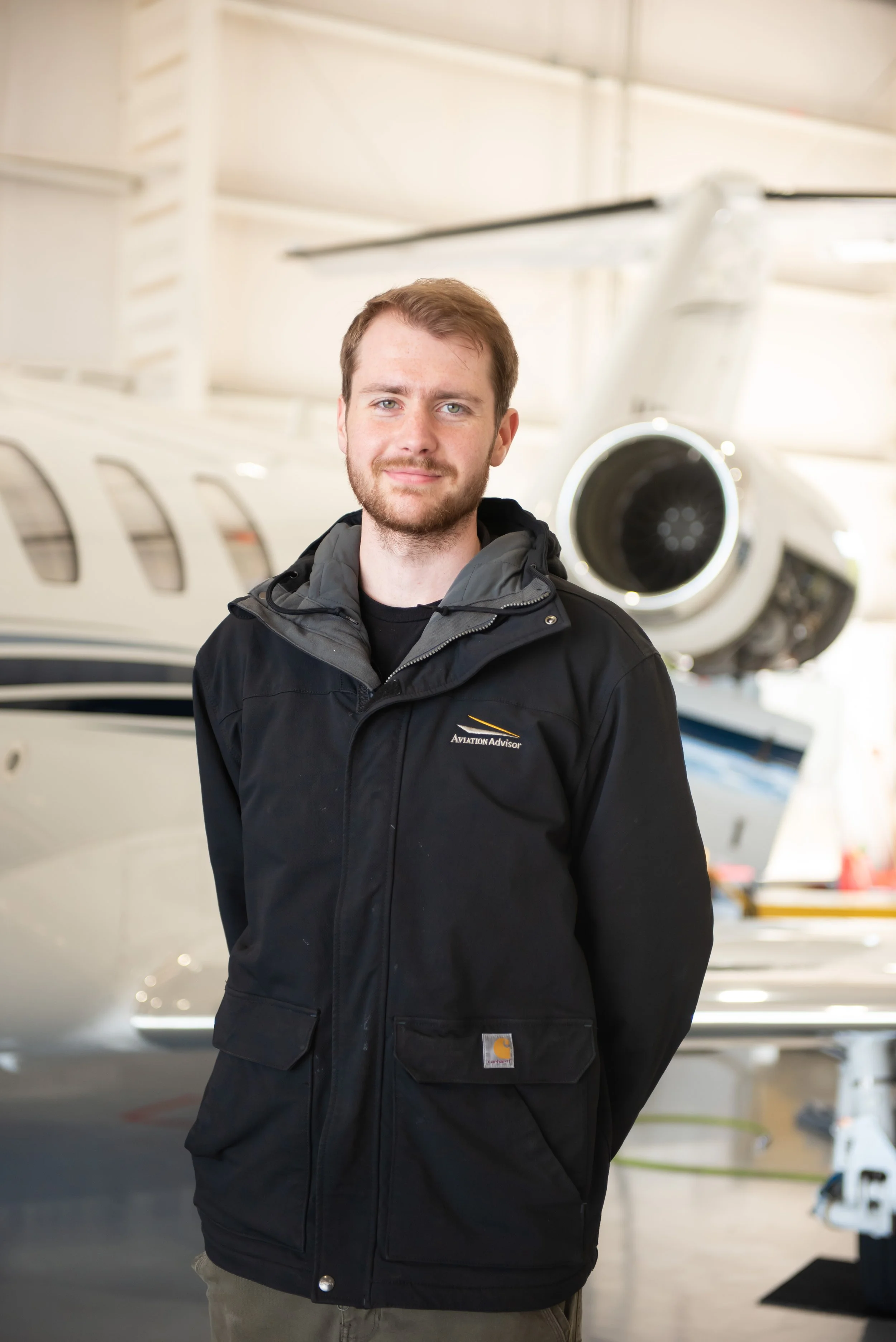 A young man with light skin, short light brown hair, and a beard standing in front of a white private jet, wearing a black jacket with a Carhartt logo and a gray hoodie underneath.