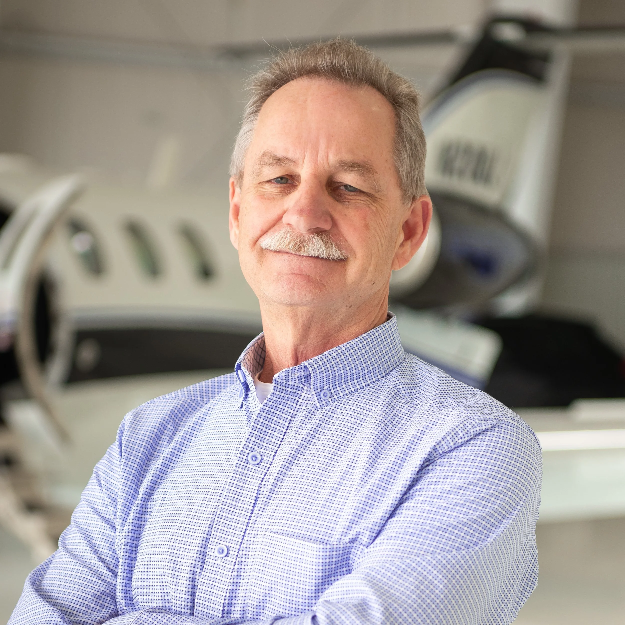 Close-up of Saul Thompson, wearing a blue and white checkered shirt, smiling in an indoor setting with aircraft in the background.
