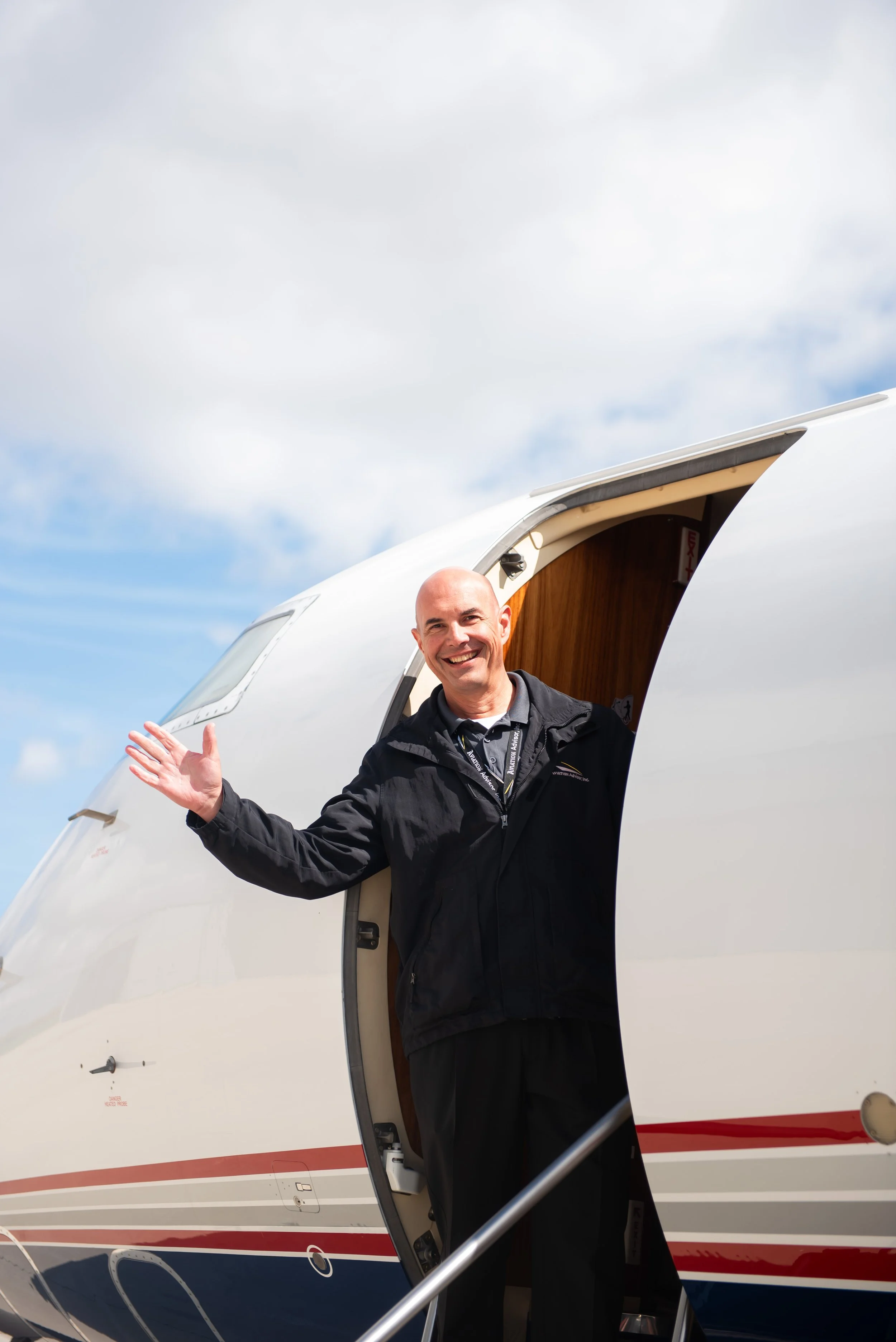 A smiling man standing at the open door of a private jet airplane, waving with his left hand, under a partly cloudy sky.