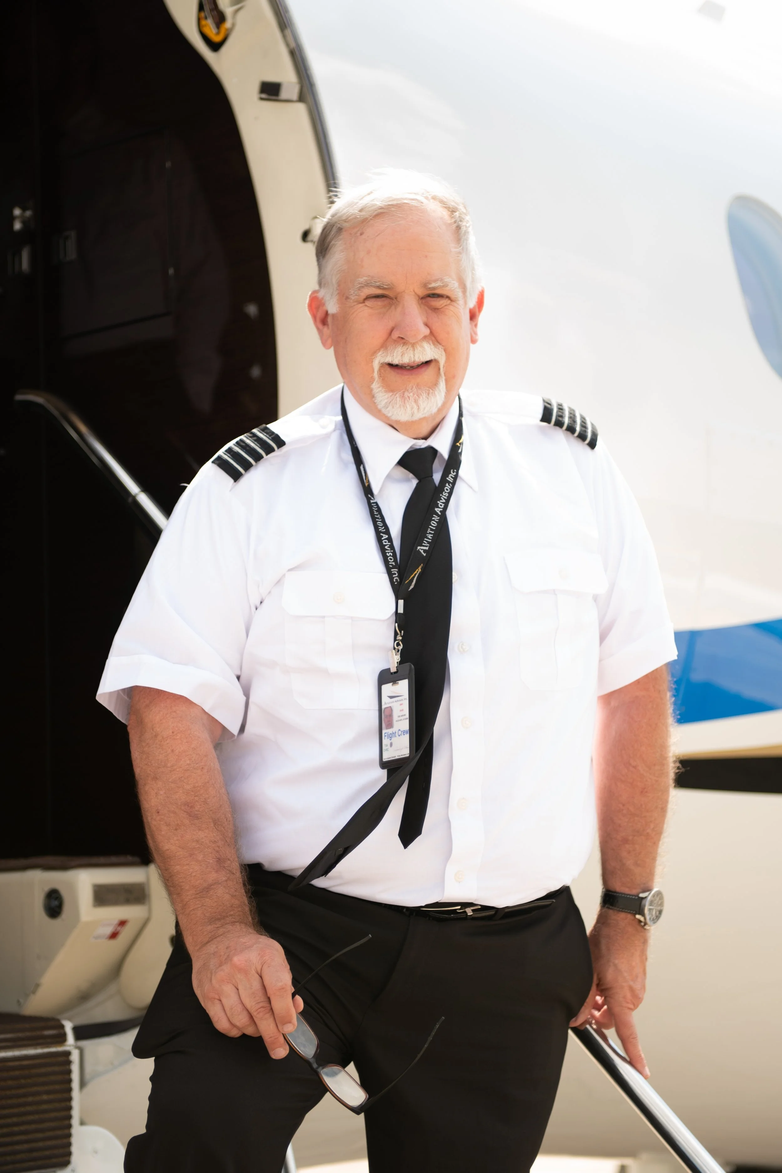 A male pilot with gray hair and a beard, wearing a white pilot uniform with shoulder epaulets, a black tie, and a lanyard with flight crew ID badge. He is holding sunglasses in his left hand and standing in front of an airplane.