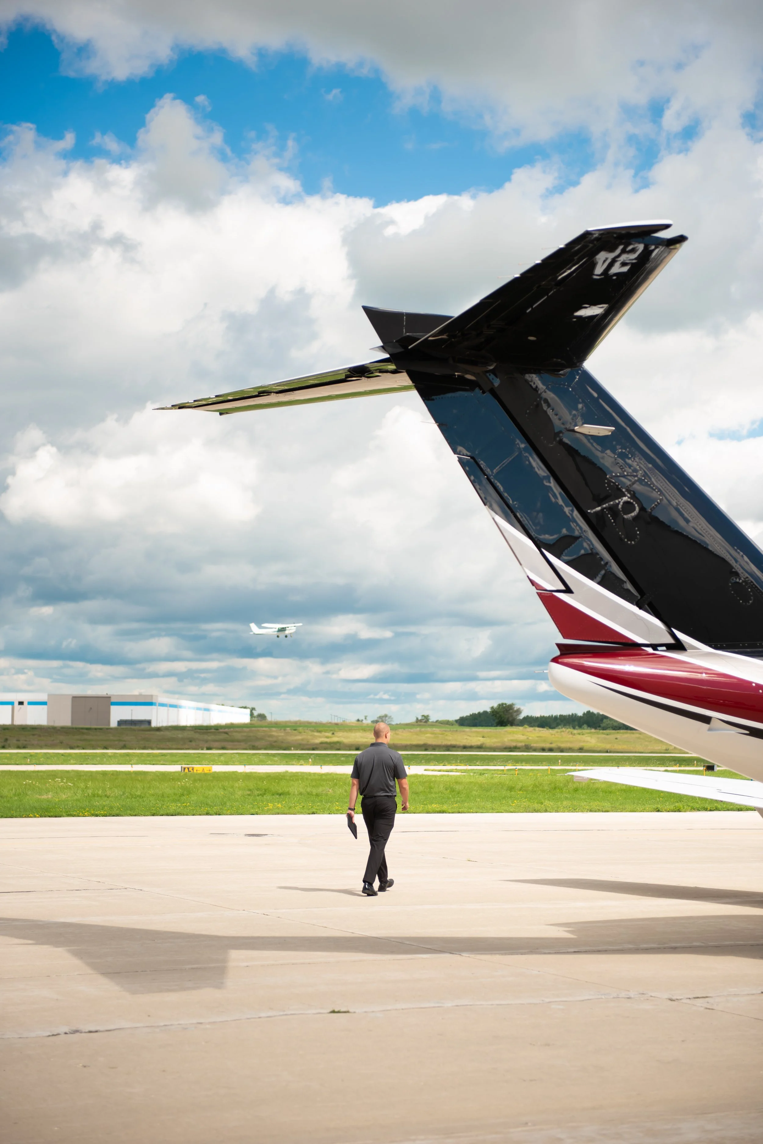 A man walking on an airport tarmac near large private jets, with a blue sky and scattered clouds above.