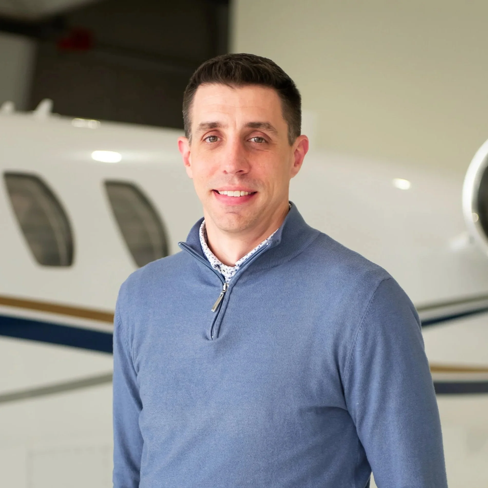 A man with short dark hair and blue eyes wearing a navy blue quarter-zip sweater over a collared shirt, standing in what appears to be an airplane hangar.