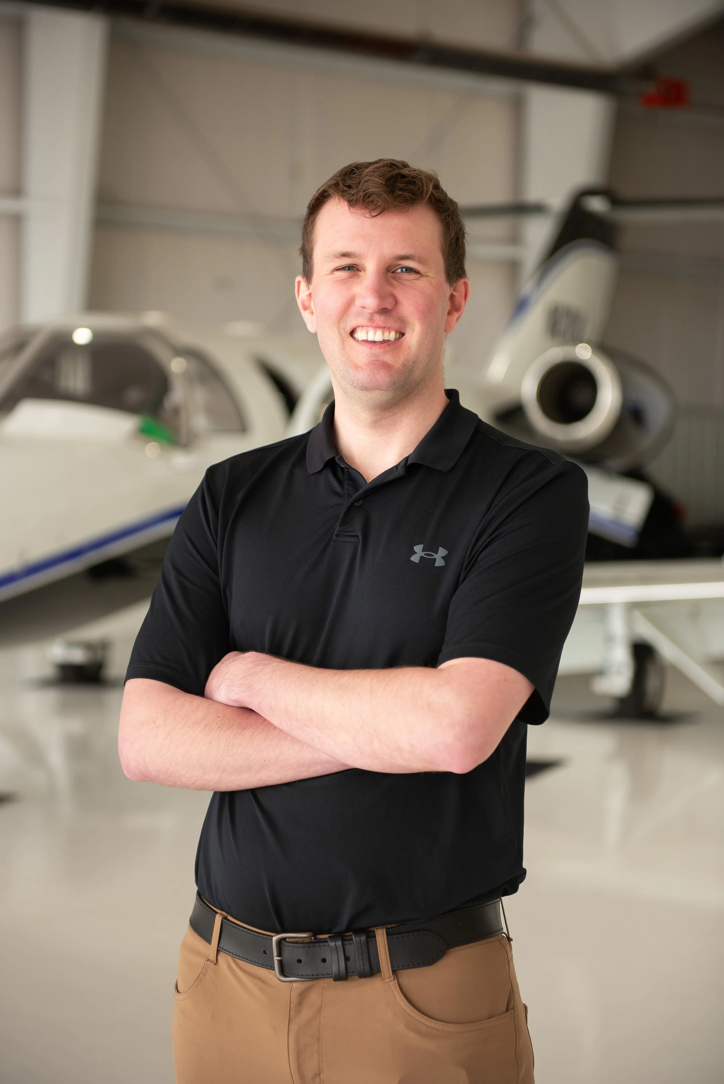 A man standing with arms crossed in front of a small airplane inside a hangar, smiling at the camera.