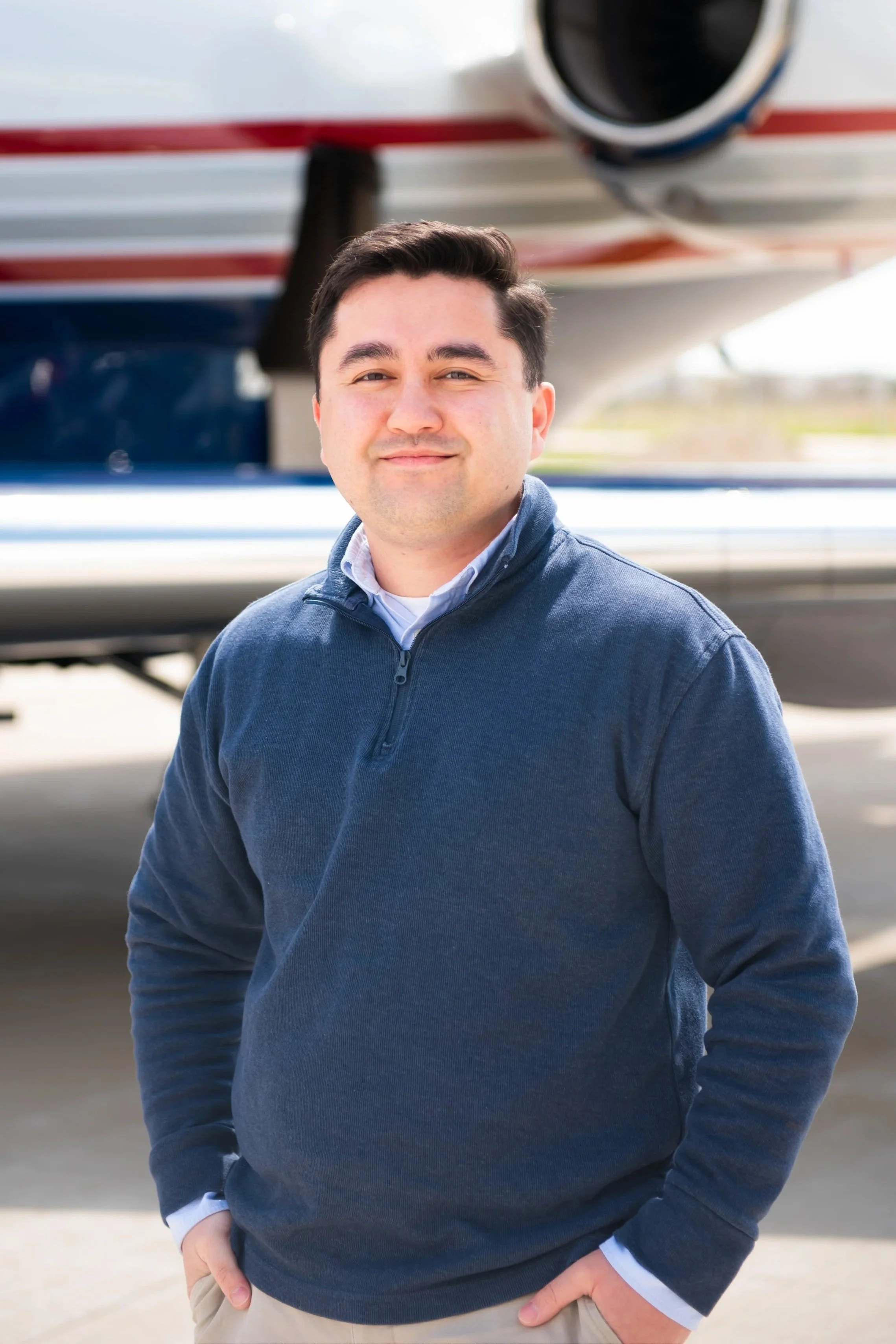 A man with dark hair, wearing a navy blue quarter-zip sweater over a white collared shirt and beige pants, standing outdoors in front of an airplane.