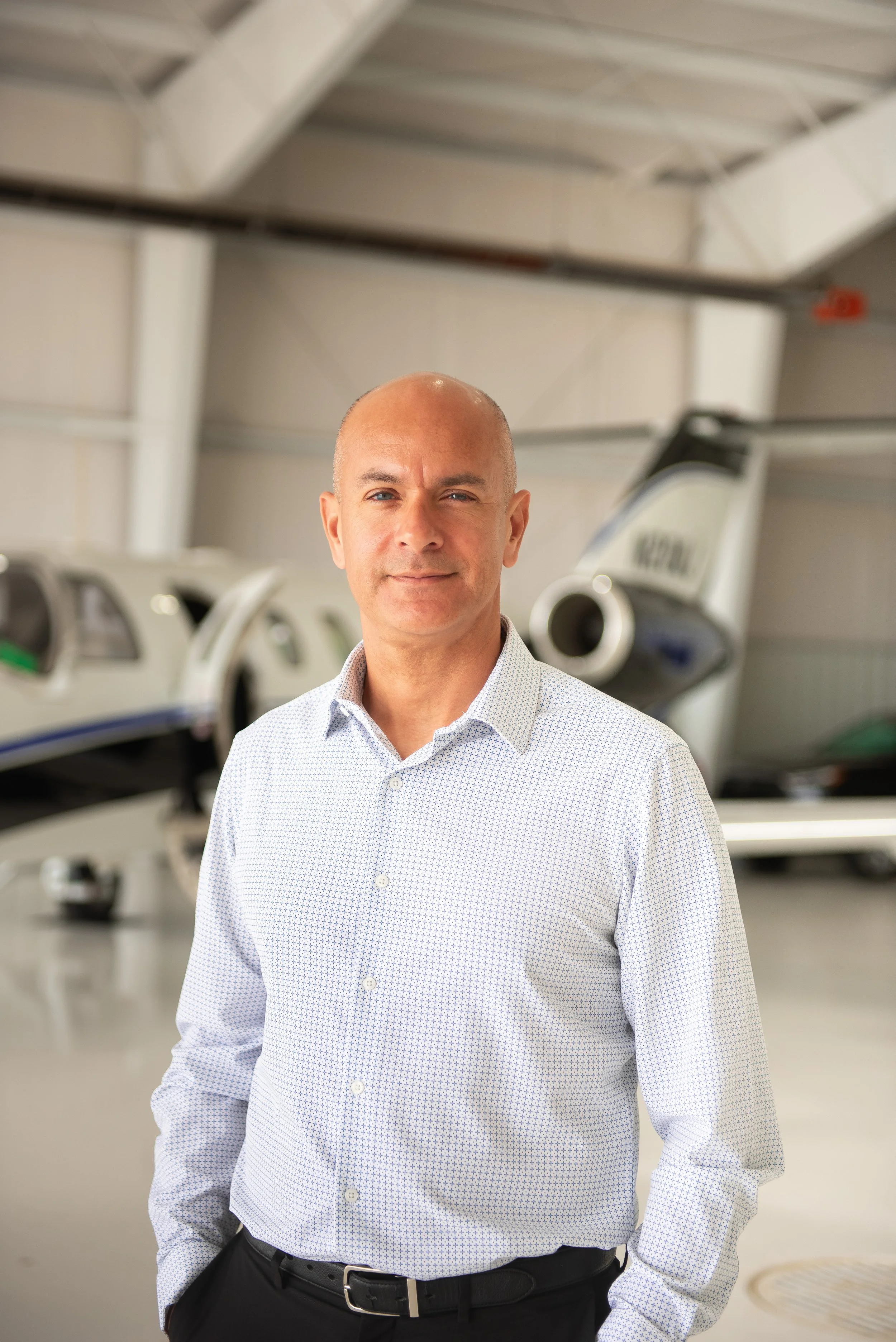 A man in a white, patterned button-down shirt standing inside a hangar with small aircraft and aviation equipment in the background.