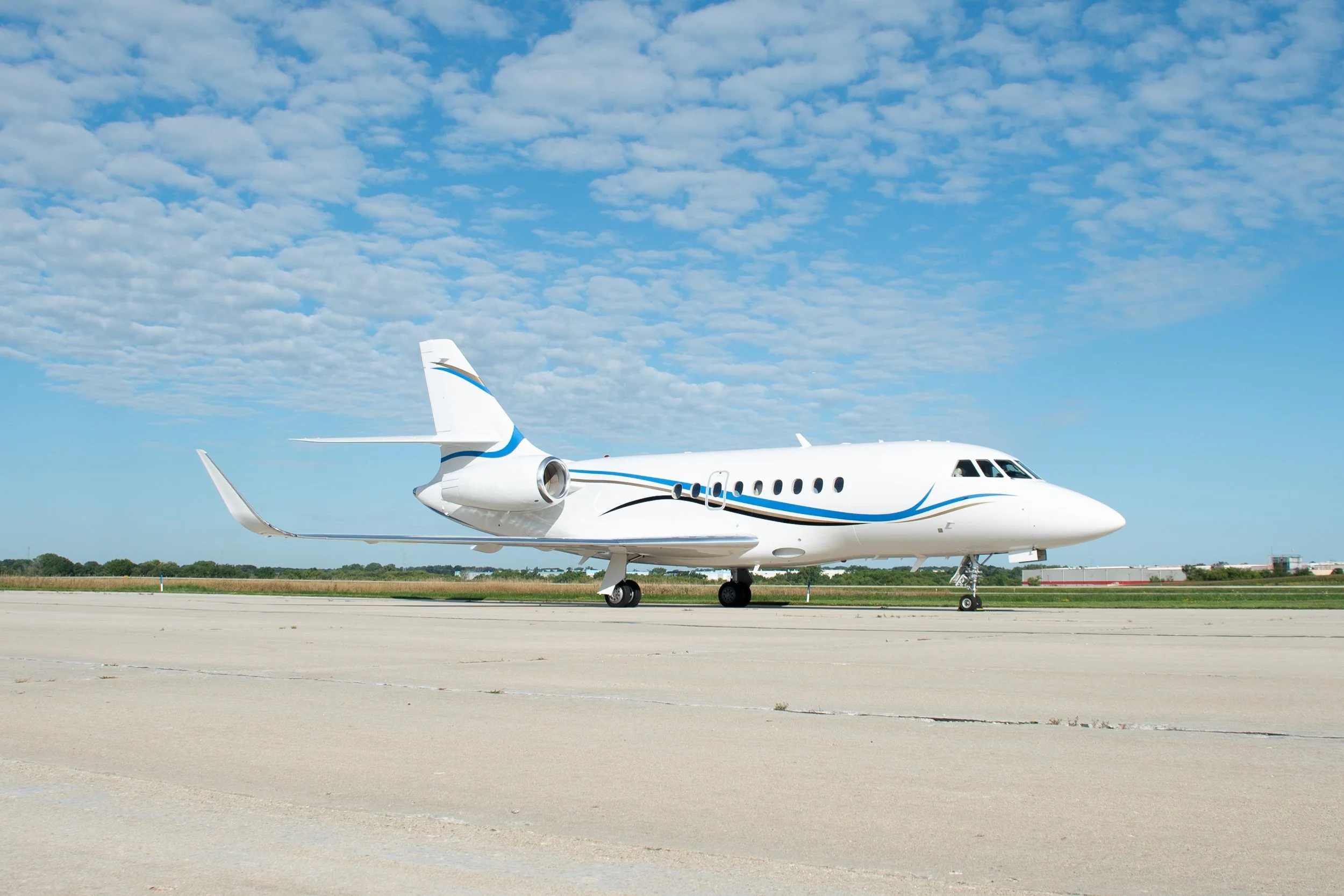 A white private jet airplane parked on an airport tarmac with blue sky and scattered clouds overhead.