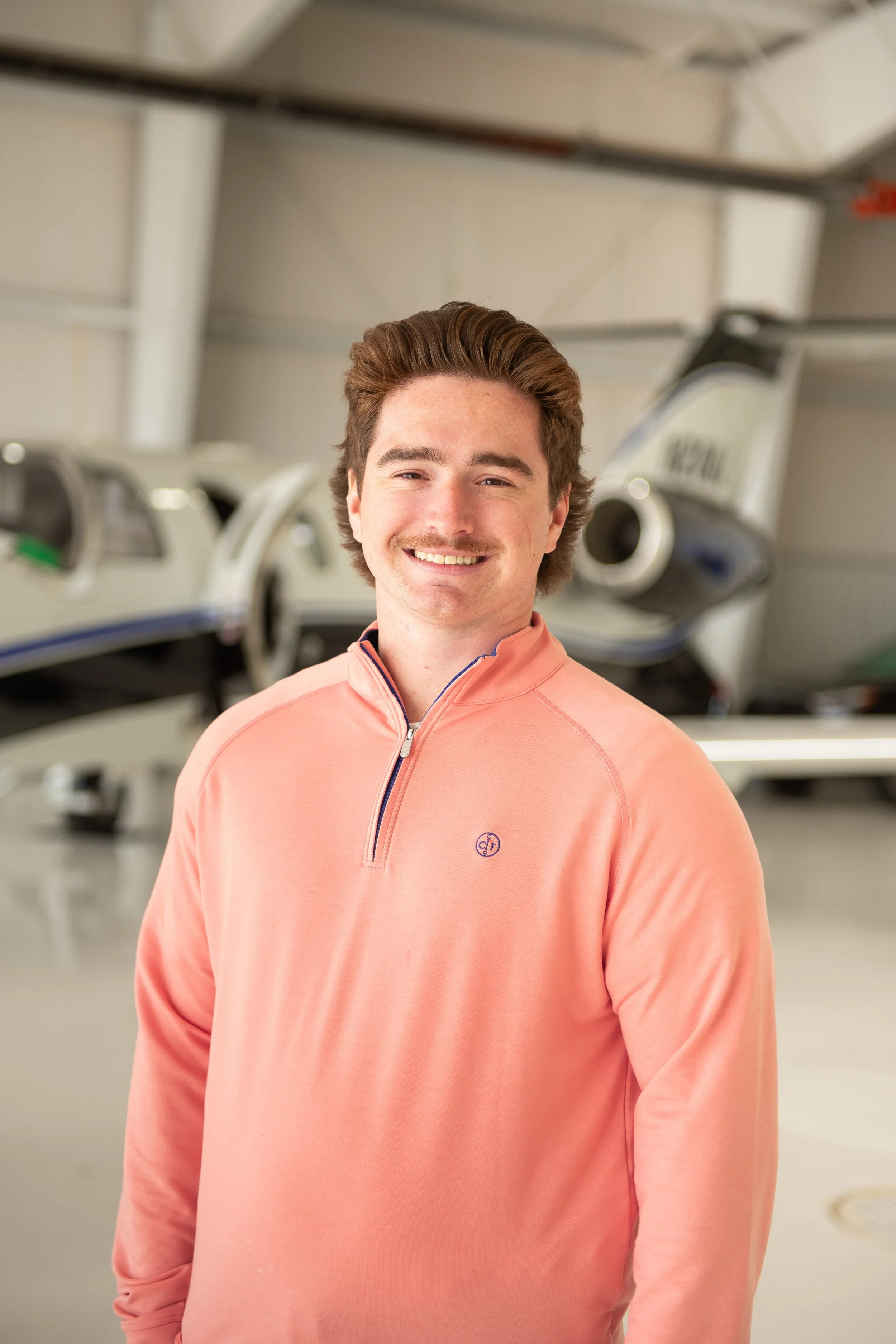 Young man with brown hair and a mustache smiling while wearing a peach-colored jacket, standing in an indoor hangar with airplanes in the background.