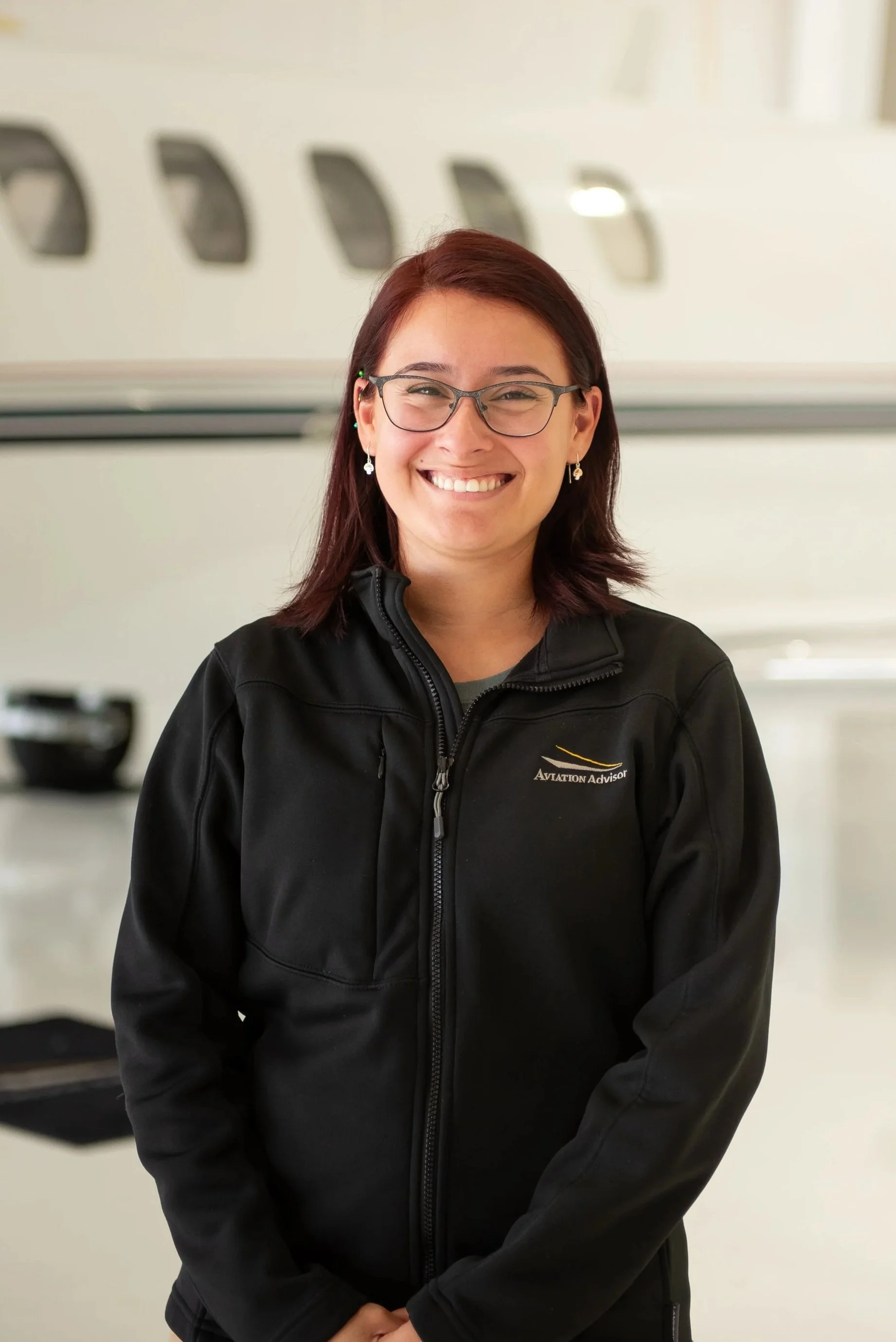 A woman with red hair, glasses, and earrings smiles at the camera, wearing a black jacket with an aviation logo, standing indoors with blurred background of an aircraft.