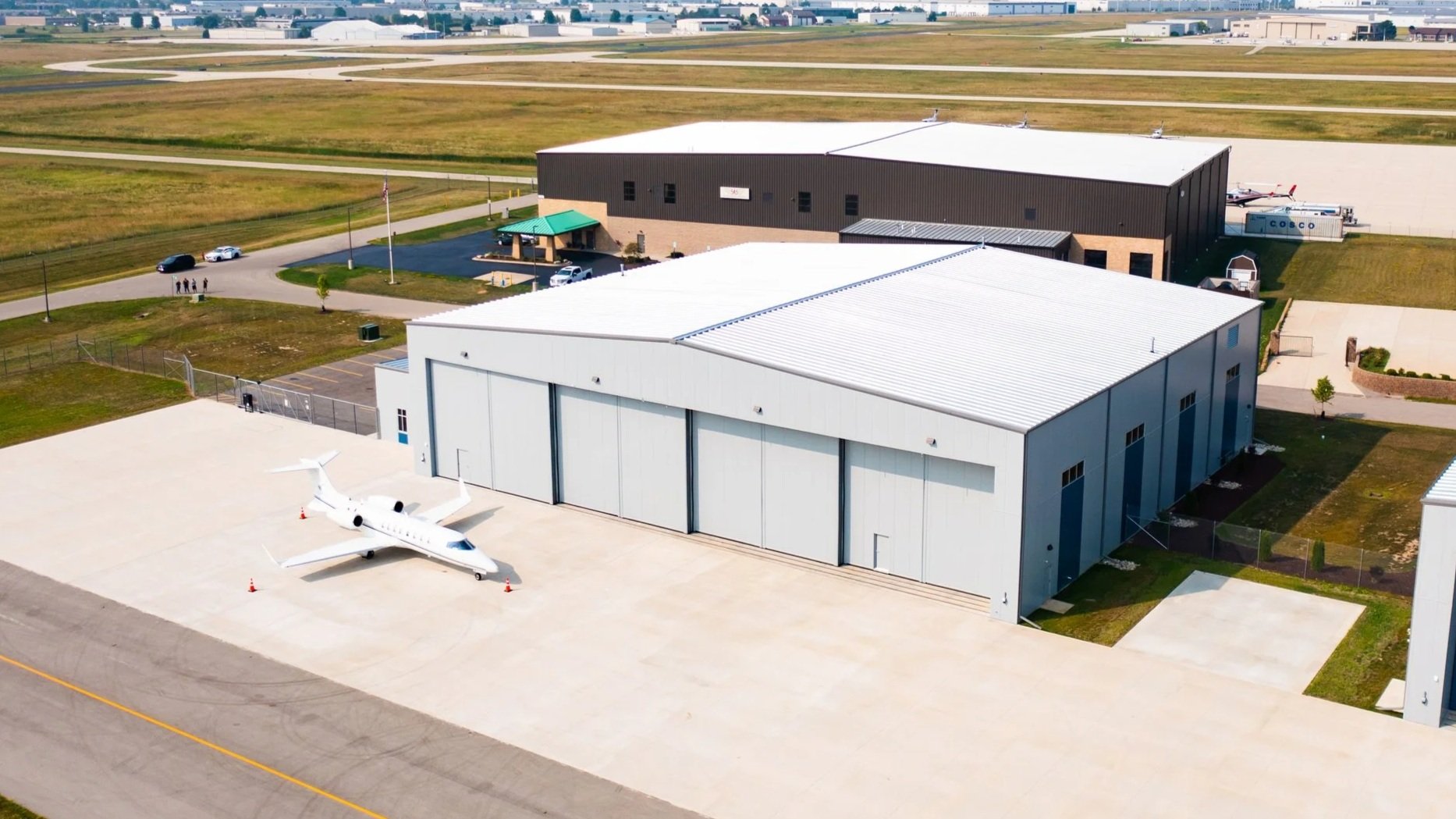 A small white private jet parked on the tarmac next to a large hangar. There are two additional hangars in the background and a small group of people near a circular driveway. Surrounding the area are open fields and airport buildings.