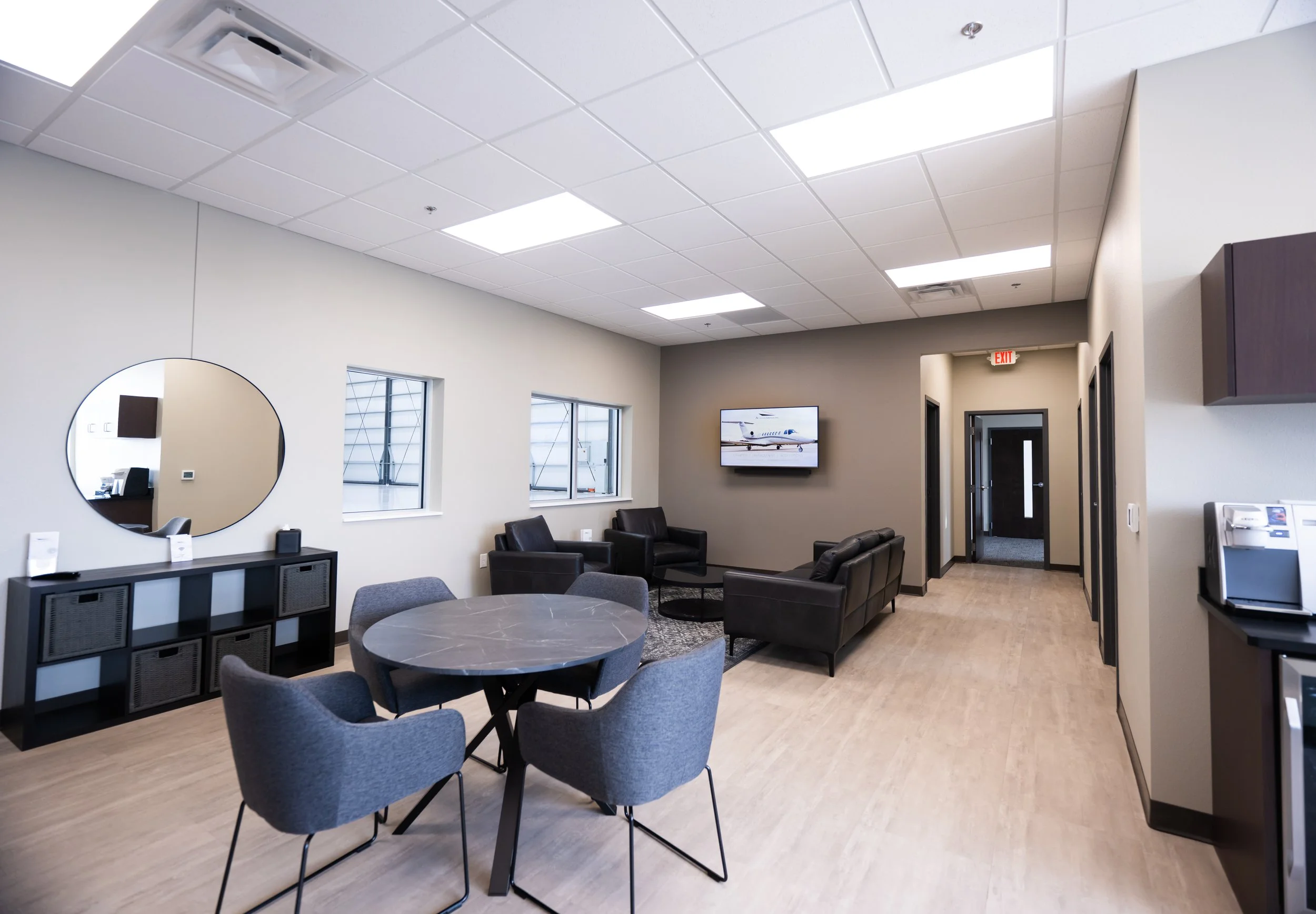 Interior view of a modern waiting area with a round table, gray upholstered chairs, a black leather sofa, a wall-mounted television displaying a private jet, a large mirror on the wall, windows showing an airport hangar, and fluorescent ceiling lights.