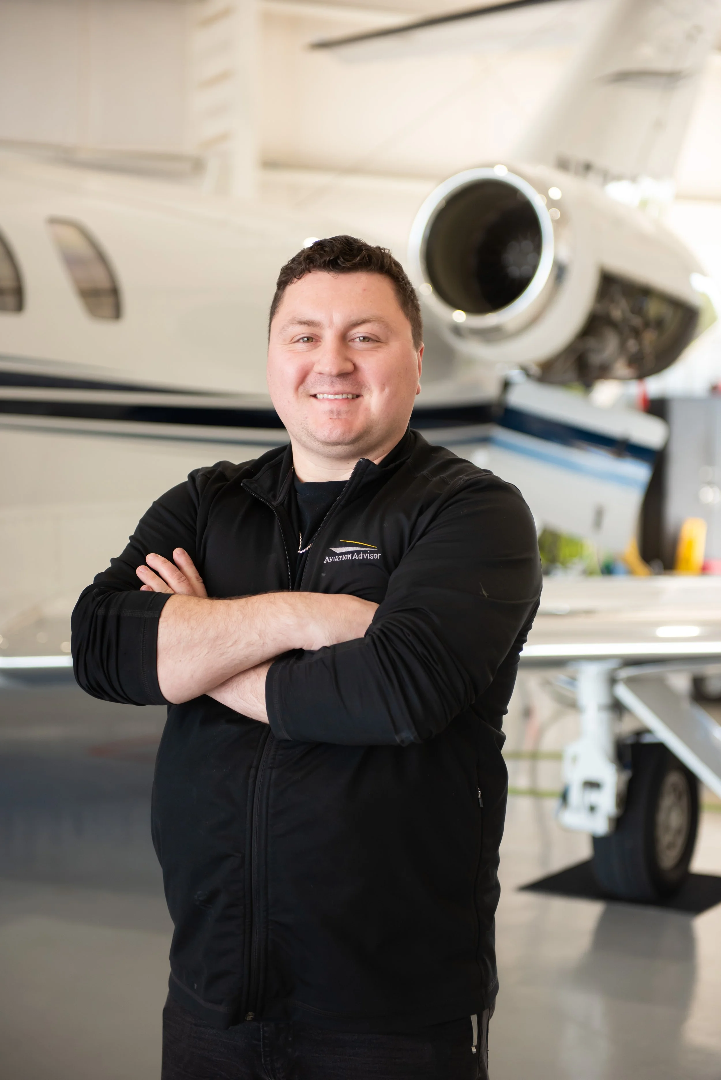 A man with short dark hair smiling and crossing his arms, standing in front of a white aircraft inside a hangar.