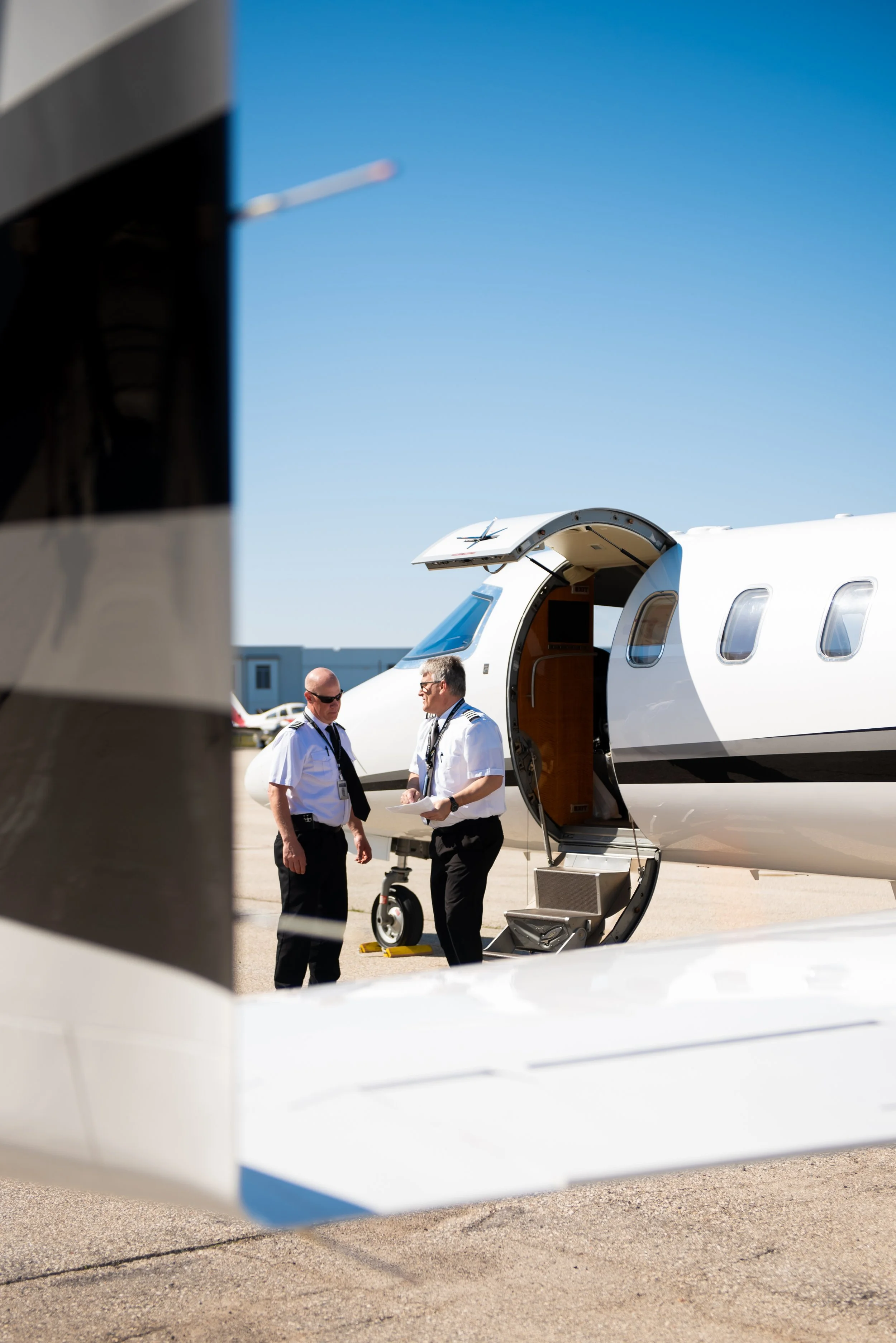 Two pilots in uniform standing outside a private jet, engaged in conversation near the aircraft's open door, with clear blue sky in the background.