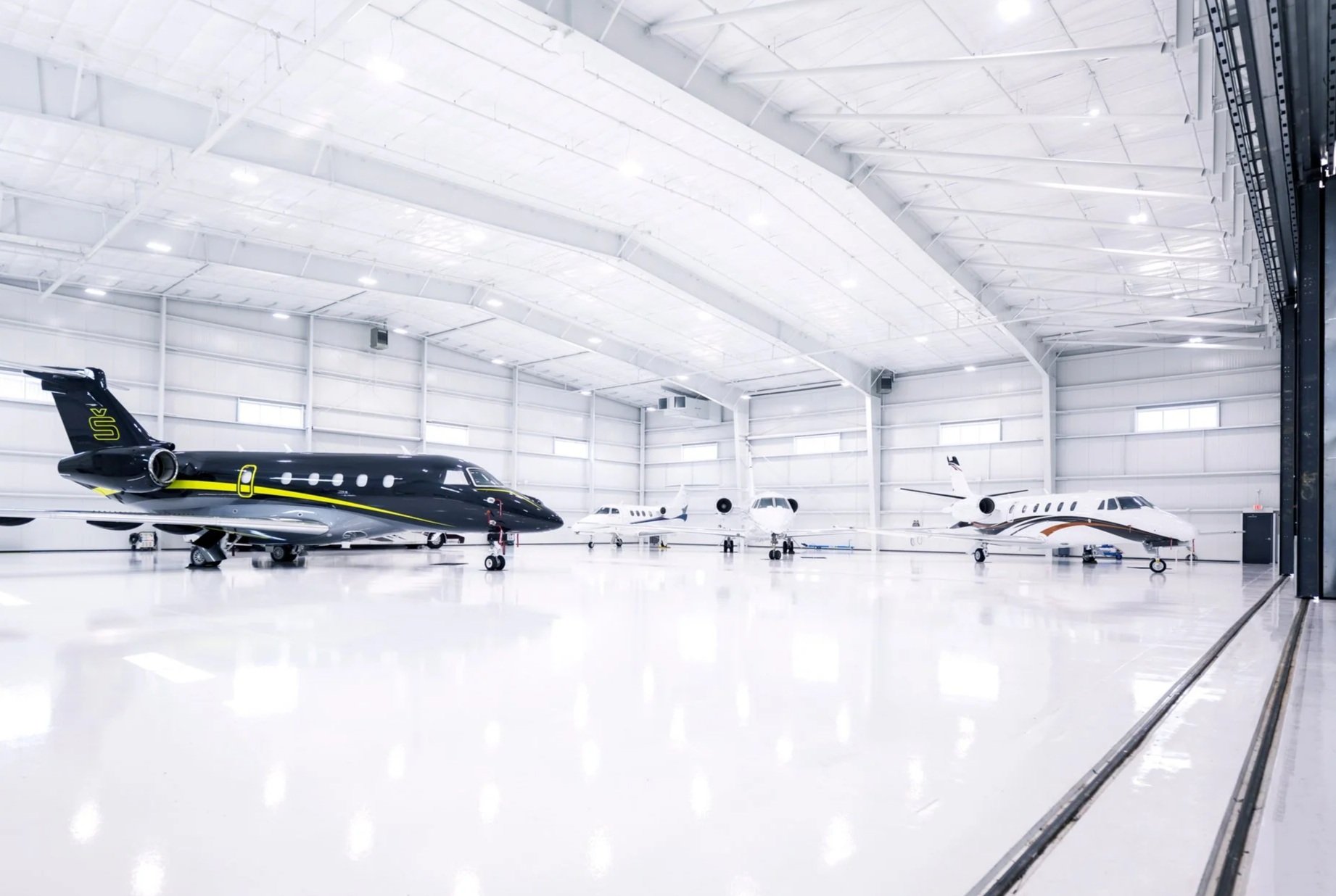 Interior of a large airplane hangar with several private jets parked inside, featuring white walls and ceiling with high, curved roof, and bright lighting.