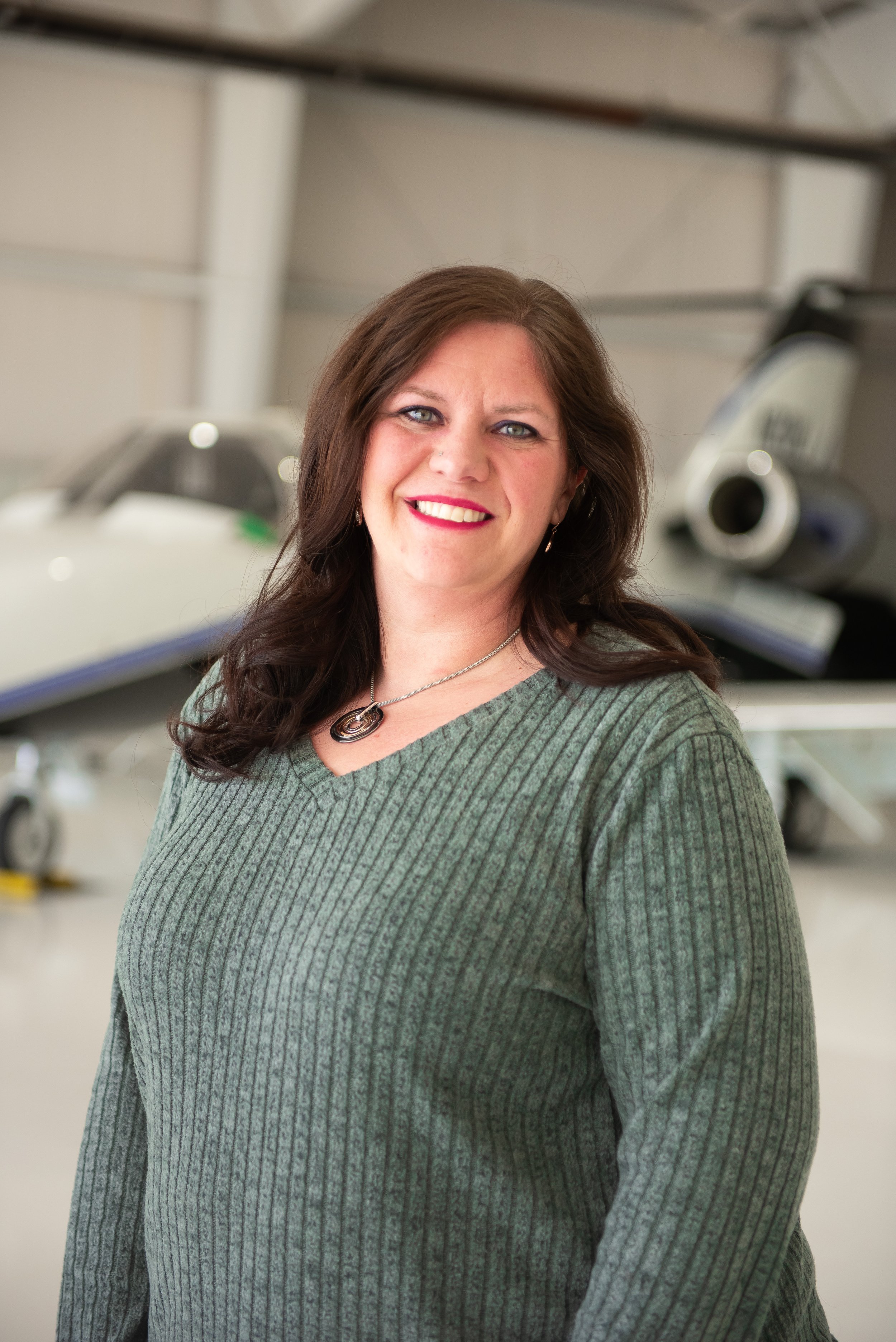 A woman with long, dark brown hair and blue eyes smiling and wearing a gray ribbed sweater standing in front of a small jet airplane inside an aircraft hangar.