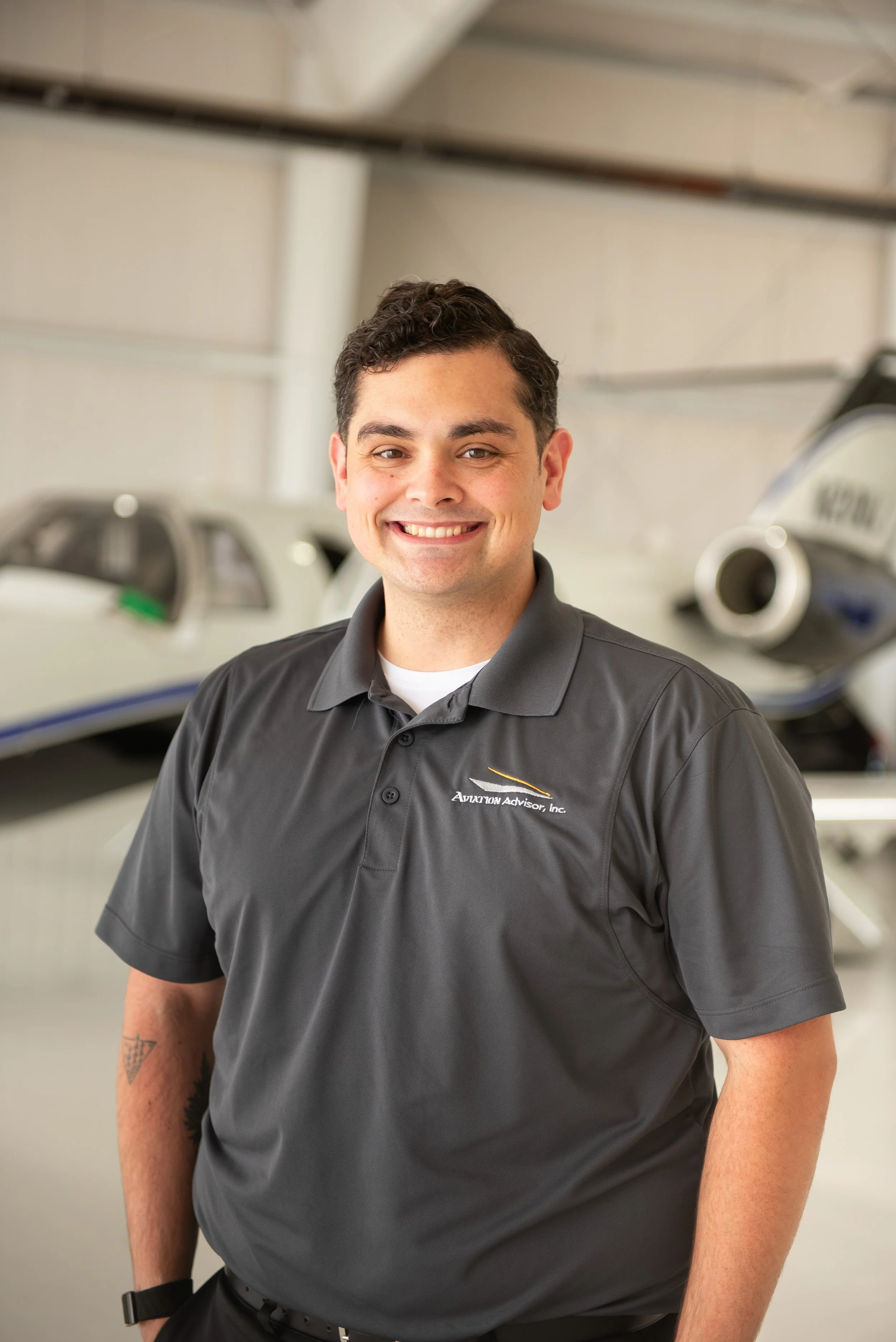 A young man with dark, curly hair and a broad smile, wearing a dark gray polo shirt with an 'Aviation Advisor, Inc.' logo, standing in front of small aircrafts in a hangar.