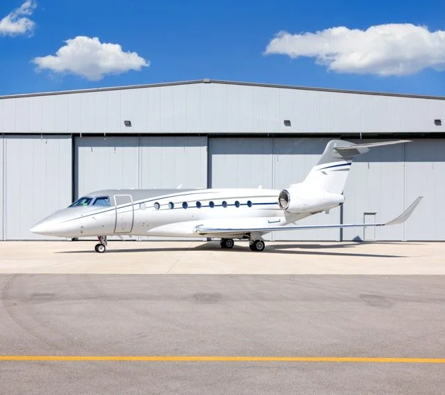 A private jet parked on the tarmac outside a large hangar on a sunny day with a partly cloudy sky.
