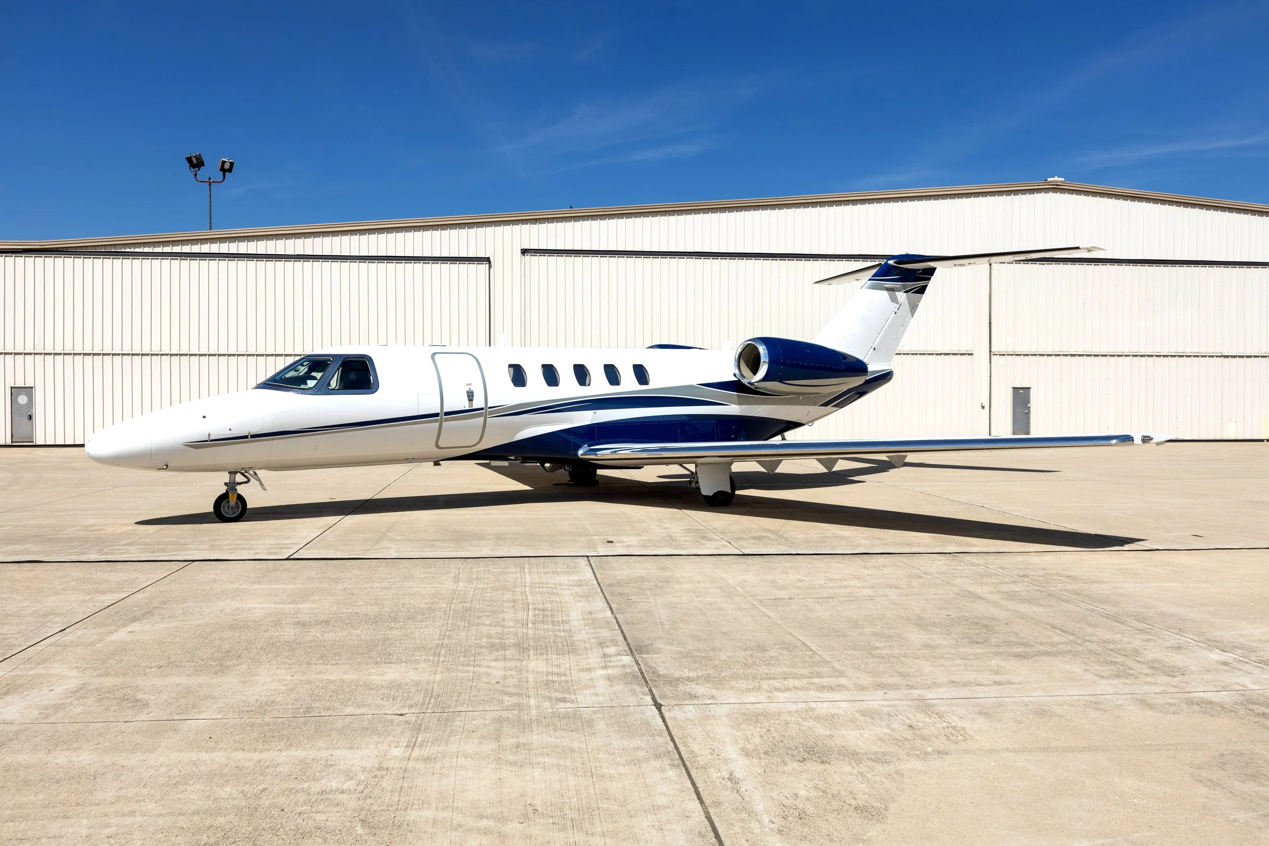 A private jet parked on the tarmac at an airport, with a beige hangar building behind it and a clear blue sky above.