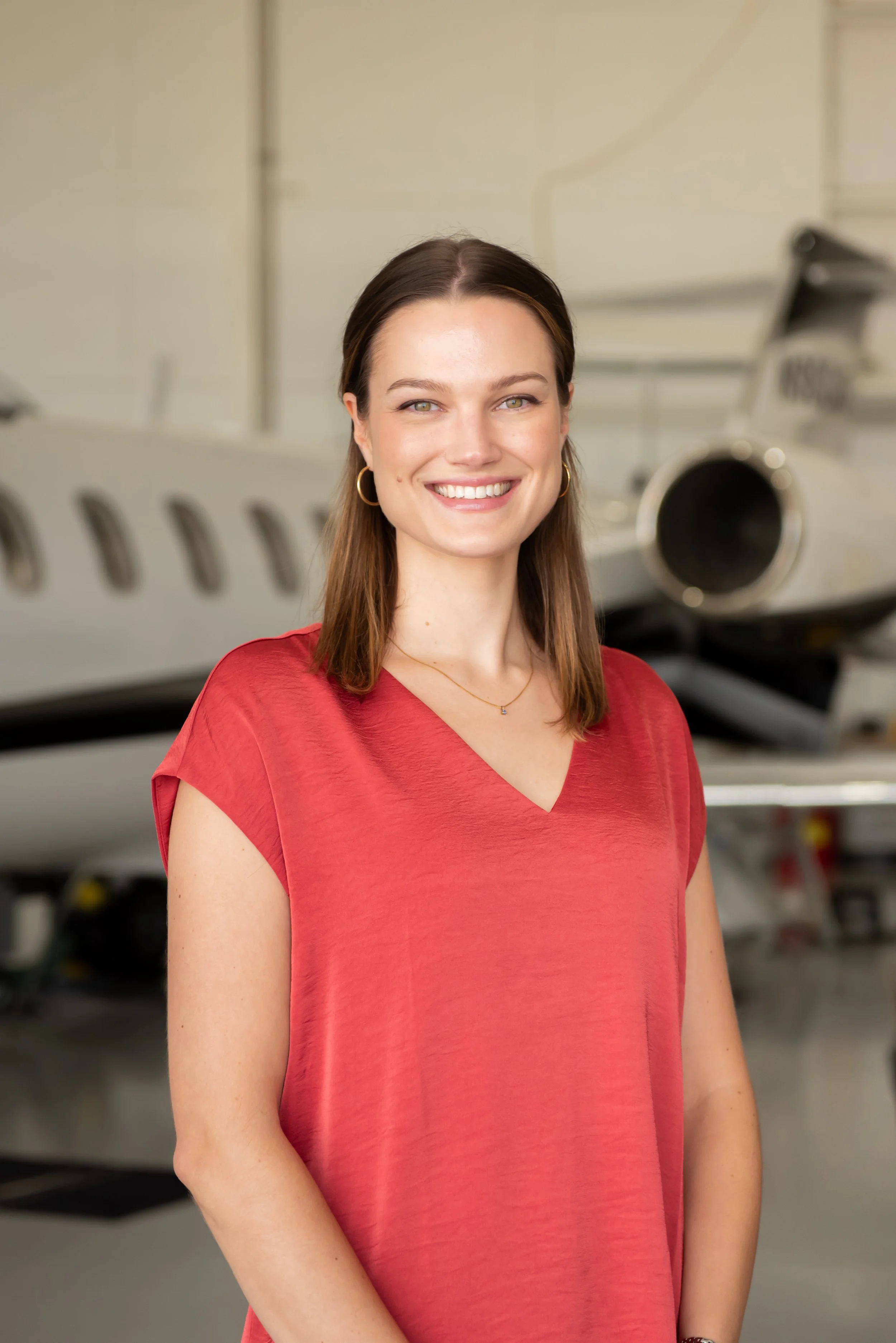 A woman with brown hair, wearing a red shirt, smiling at the camera with a background of an aircraft.