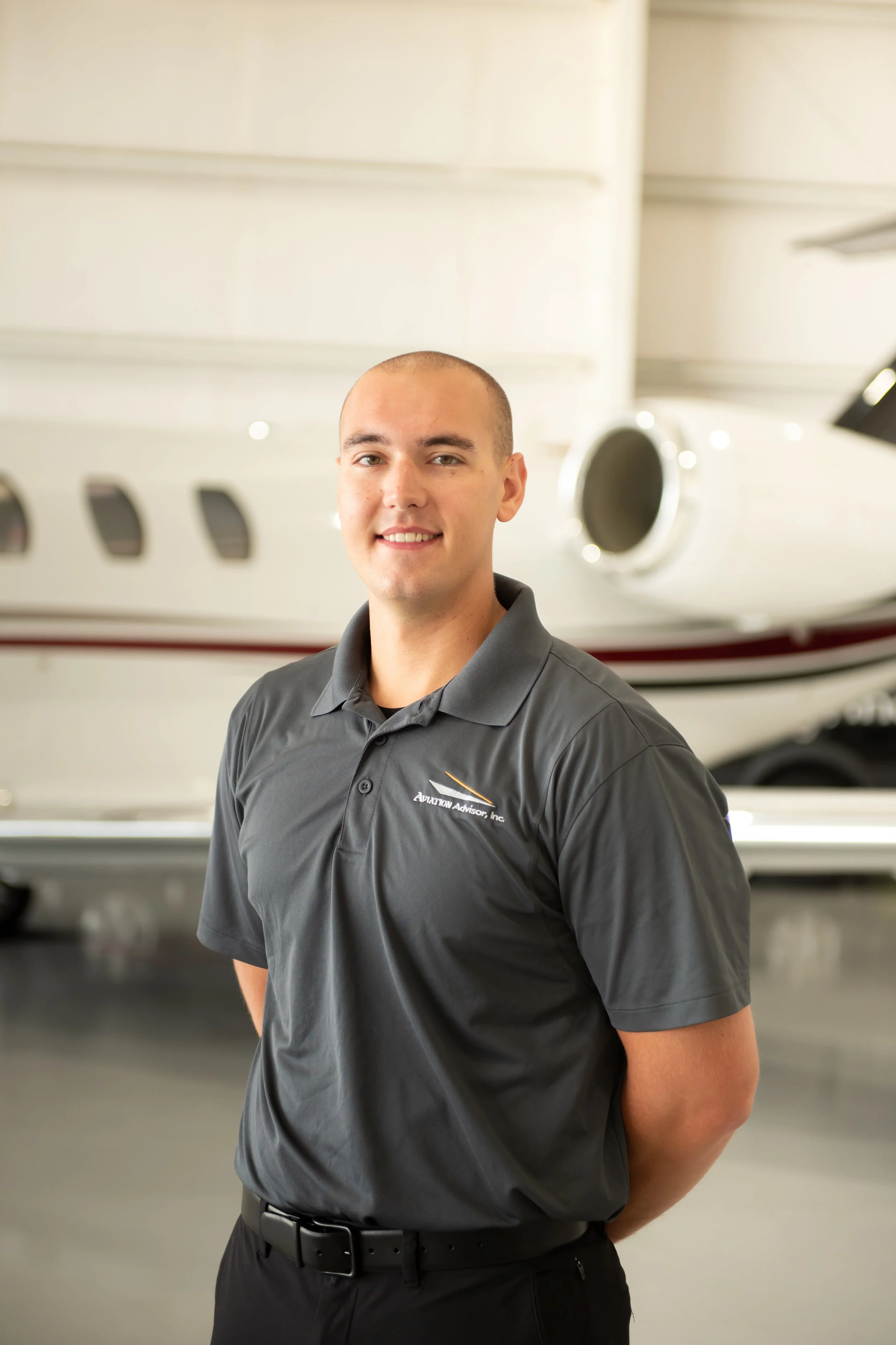 A young man with a shaved head and light skin is smiling and standing in front of a private jet inside an airport hangar. He is wearing a dark gray polo shirt with a logo that says "Aviation Advisor, Inc." and has his hands behind his back.
