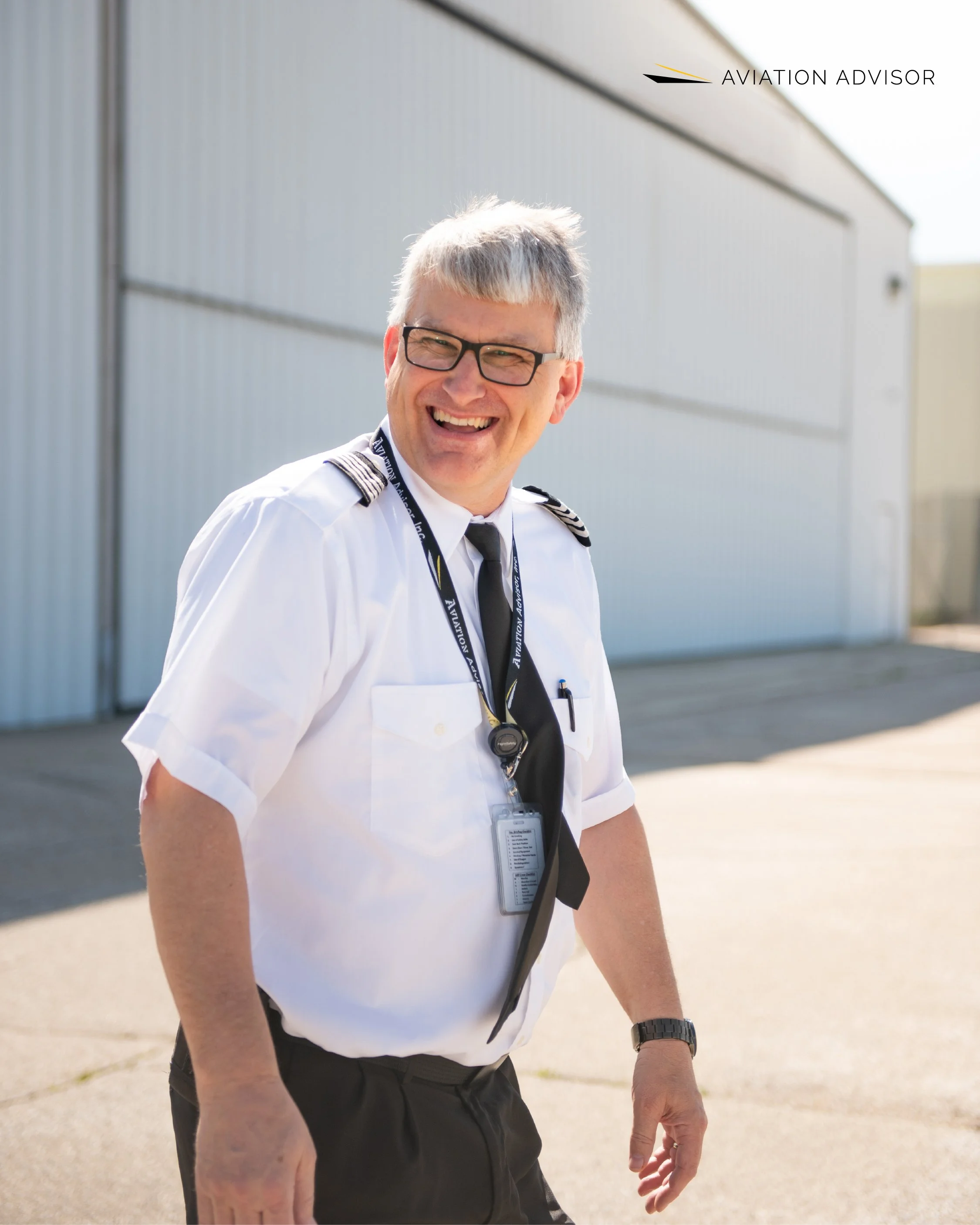 A smiling male aviation professional wearing a pilot's uniform, glasses, and a lanyard with an ID badge, standing outside near an aircraft hangar.