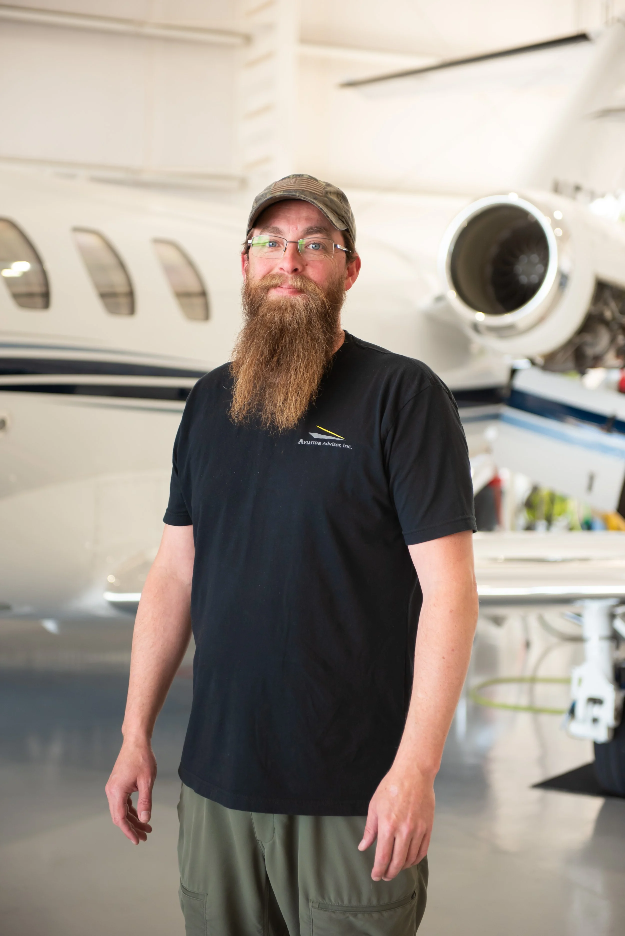 A man with a long beard and glasses wearing a camouflage hat and black T-shirt stands in front of a private jet in a hangar.