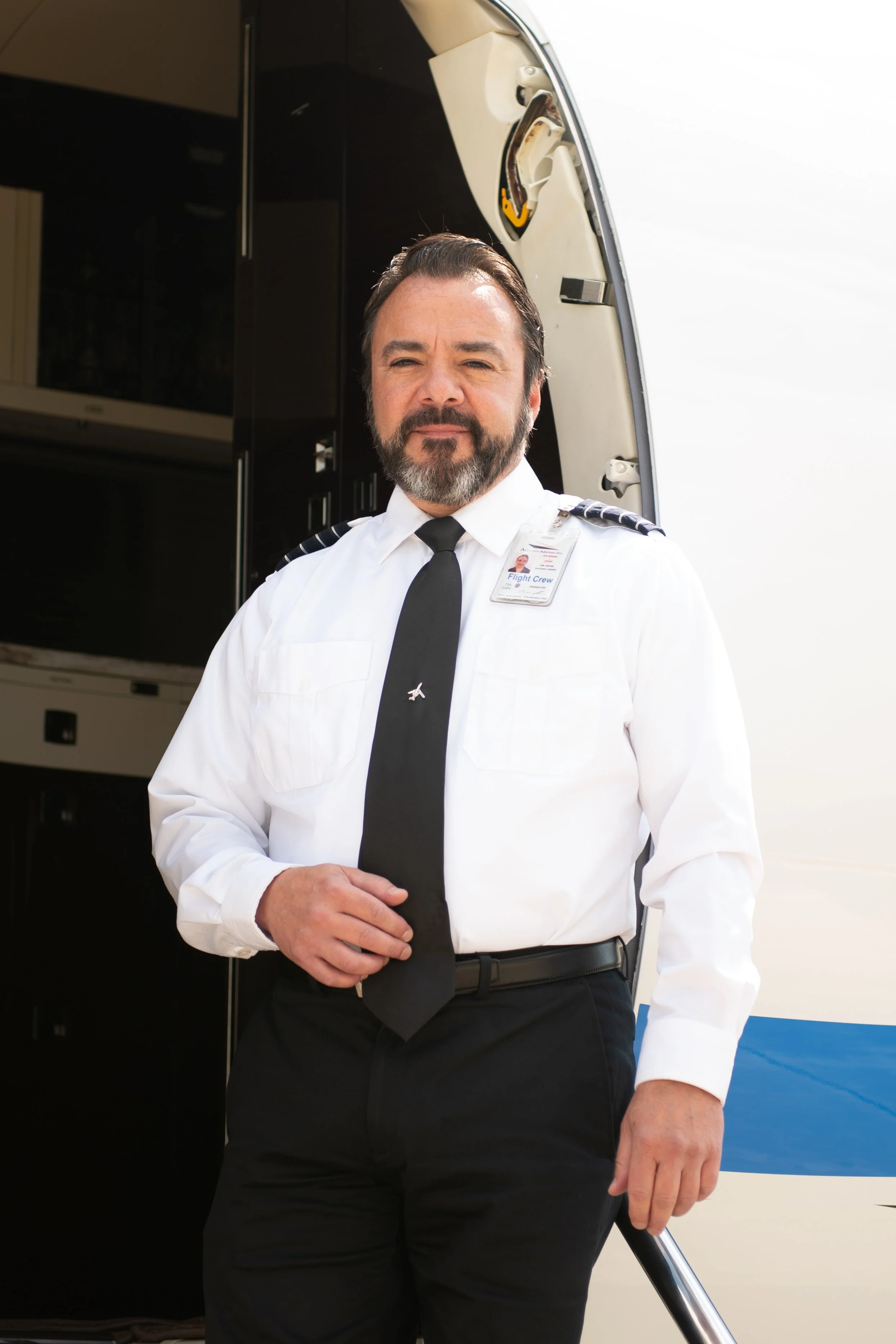 A male flight crew member dressed in a white shirt, black tie, and black pants, standing in front of an aircraft door. He has a beard and is wearing a flight crew ID badge.