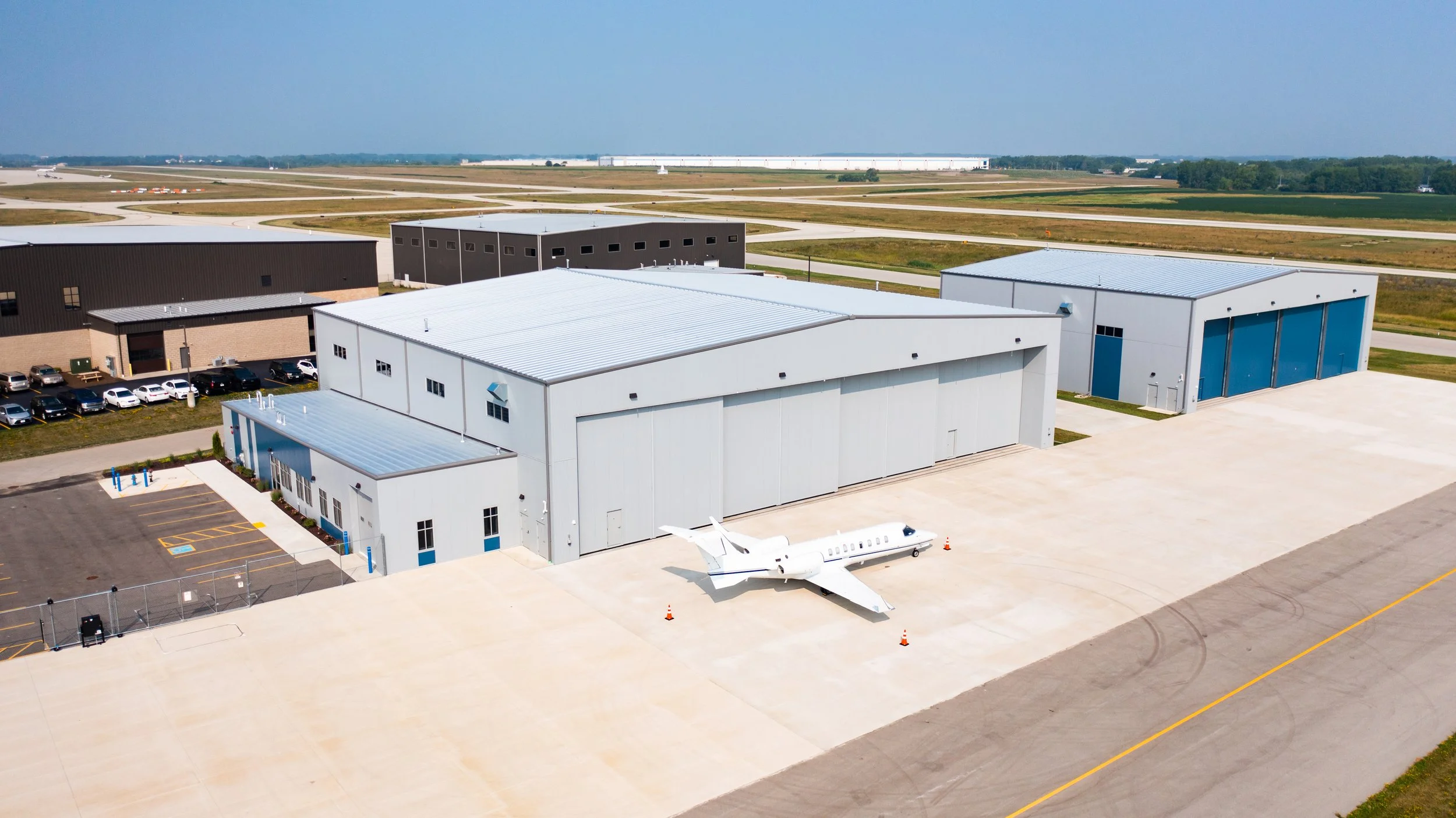 Aerial view of small airport terminal with private jet and surrounding hangars, runway, and open fields.