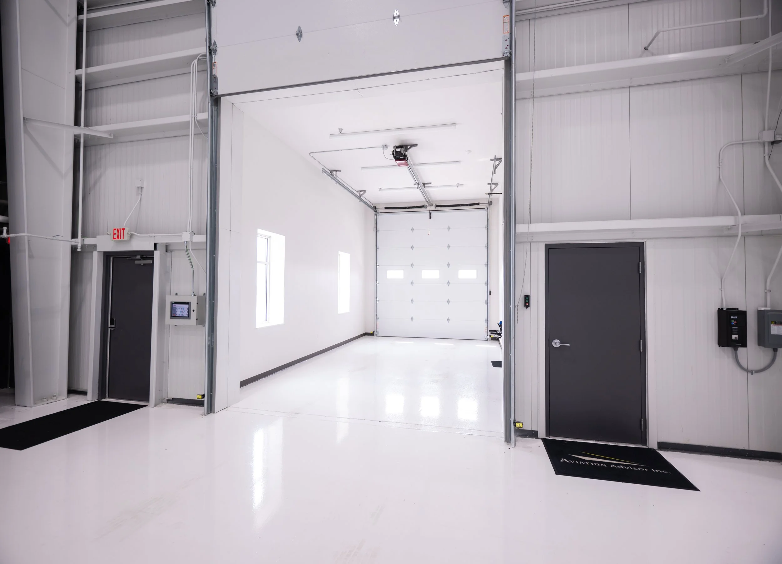 An indoor facility with white walls and floors, featuring a large garage door, smaller door, and equipment mounted on the walls. The space appears clean and brightly lit.