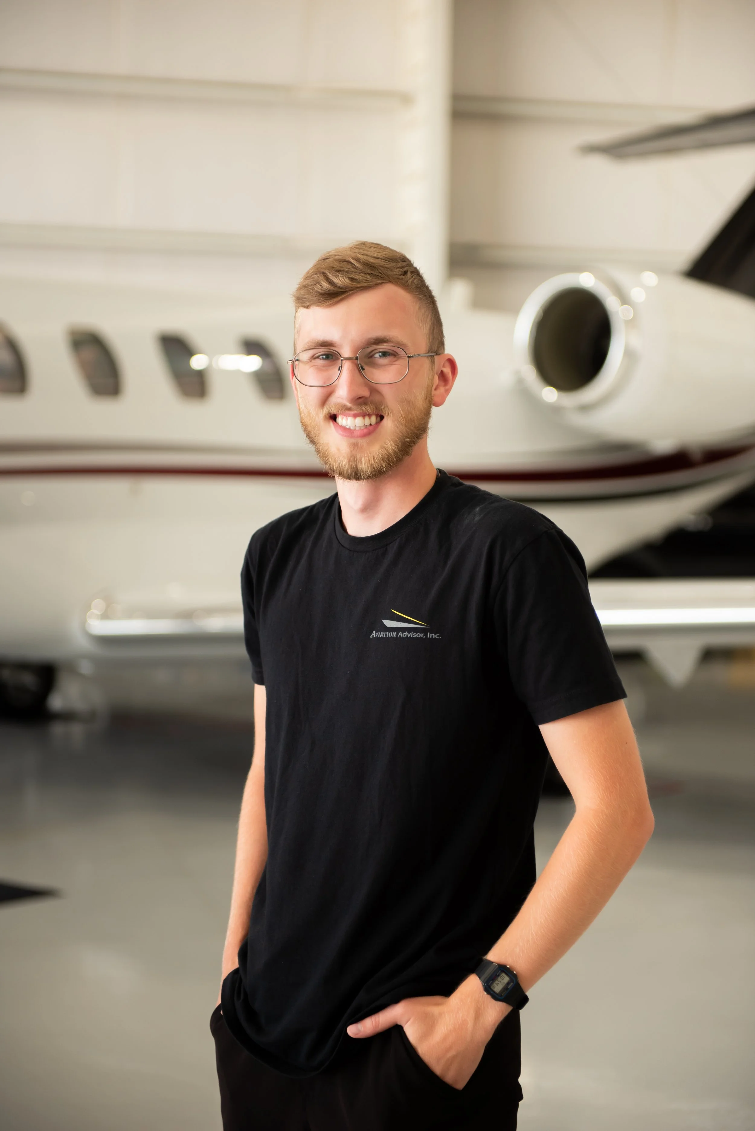 Young man with glasses and a beard smiling, wearing a black T-shirt with a logo, standing in front of a private jet in hangar, with his hands in his pockets.