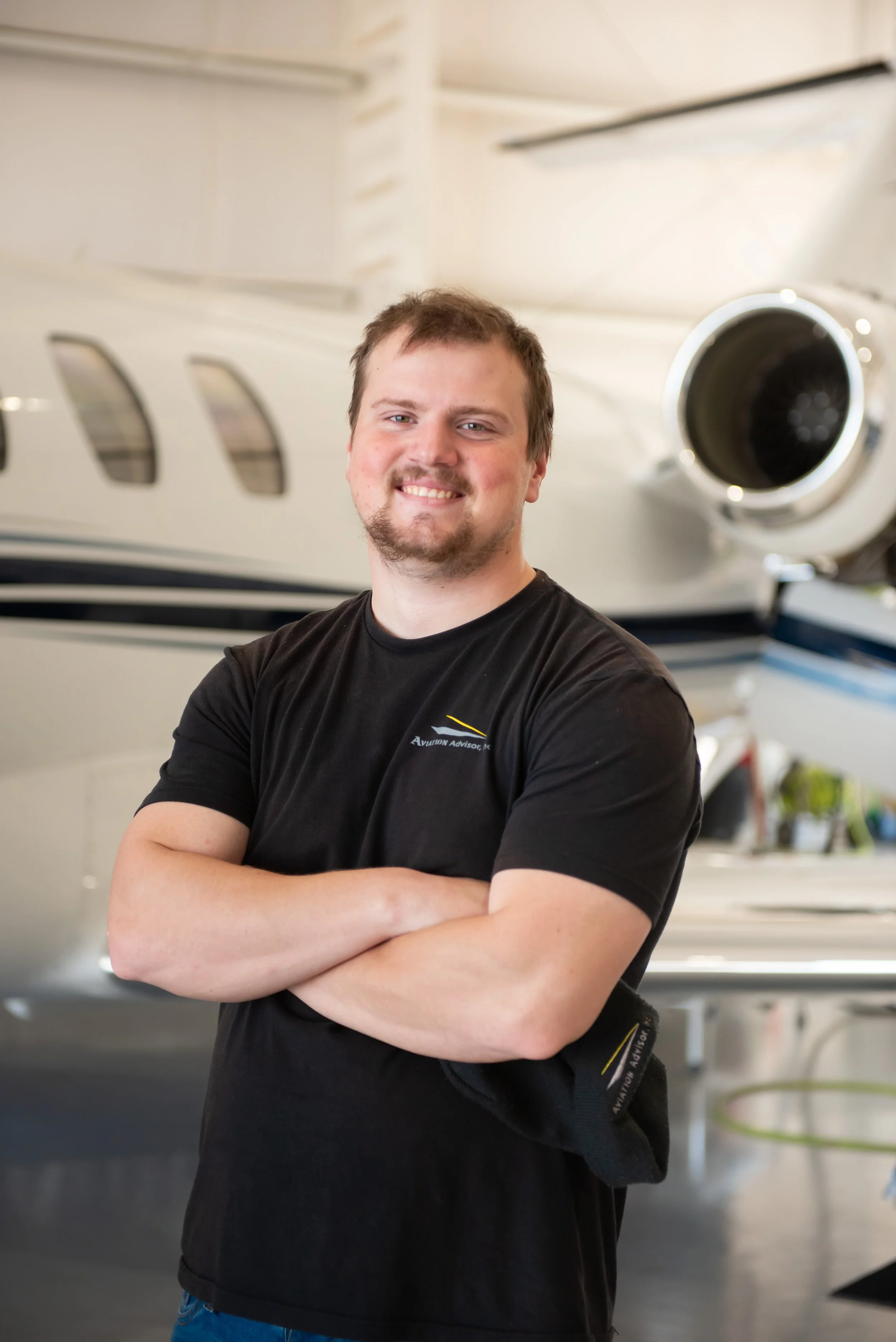 Man standing with arms crossed in front of a large aircraft engine, smiling, in an indoor hangar.