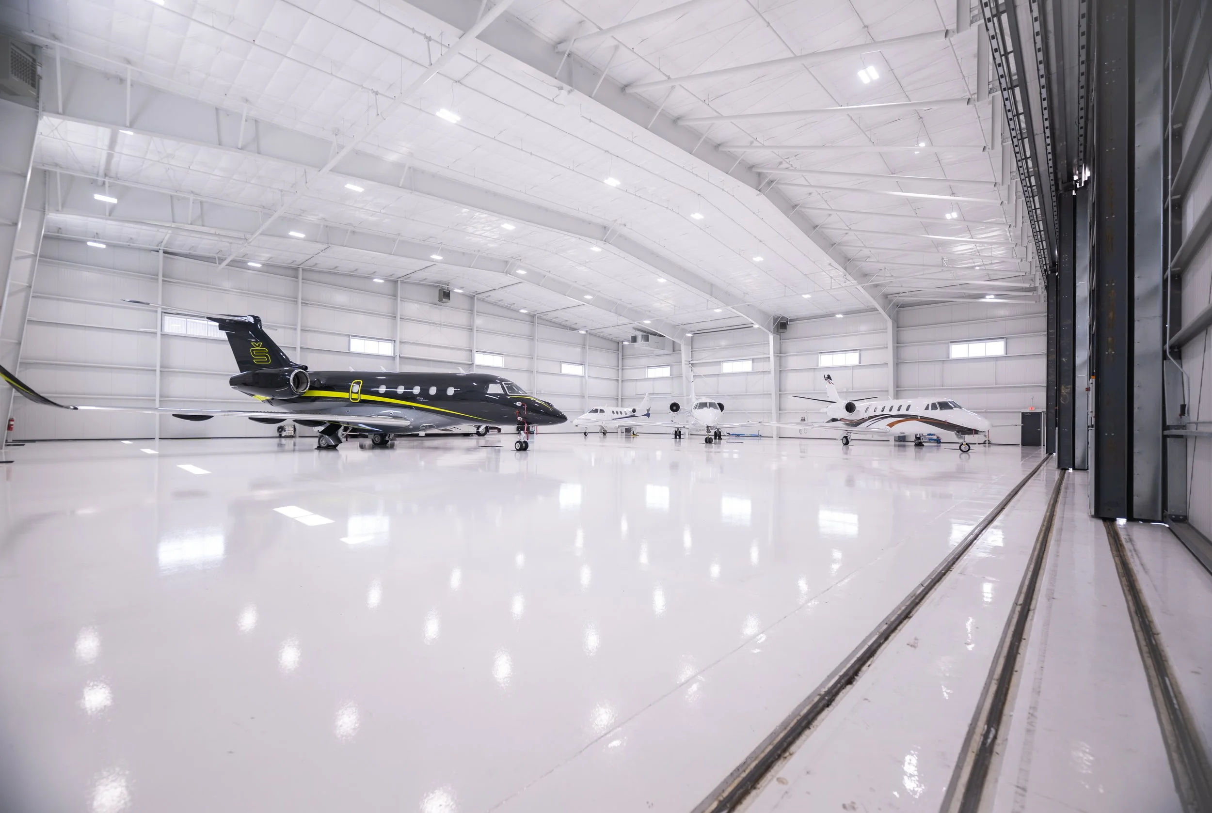 Interior of a private jet hangar with multiple private jets parked inside, high white ceiling with lighting, and a glossy white floor.