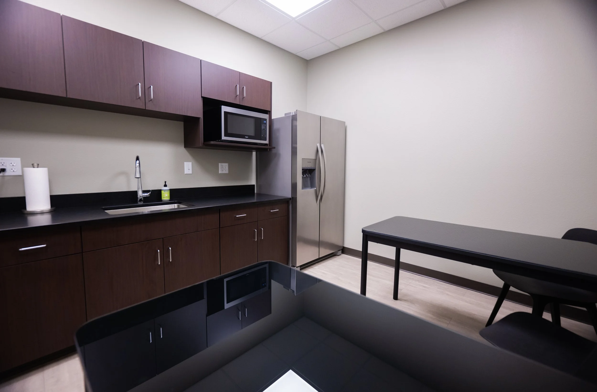 Kitchen area with dark wooden cabinets, a microwave oven, a stainless steel refrigerator, a black countertop with a sink, paper towels, and a bottle of dish soap, next to a small table and chairs.
