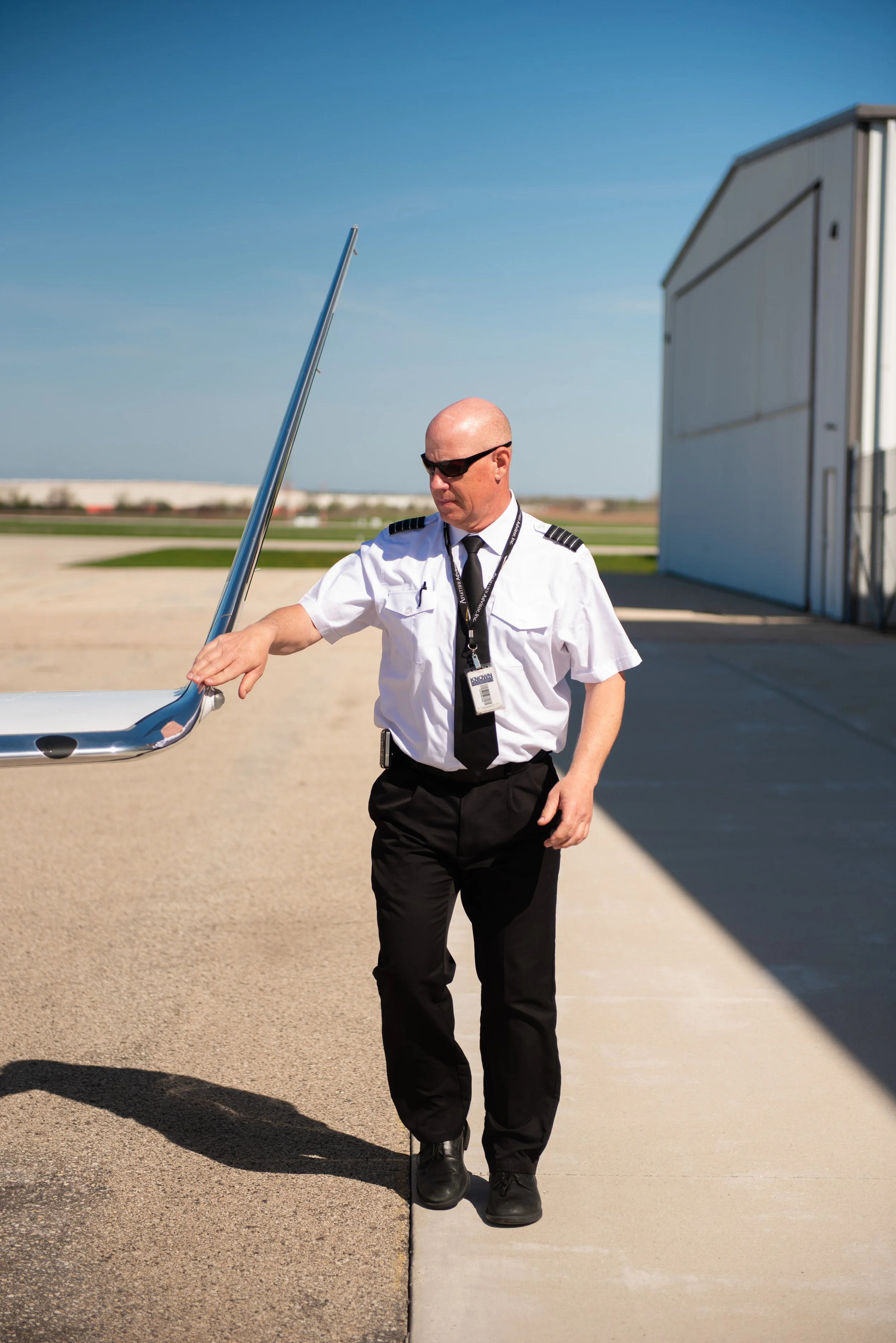 A bald man wearing security or airline staff uniform, sunglasses, black pants, and black boots, walking on a tarmac near a building while holding a metal rod or pole.