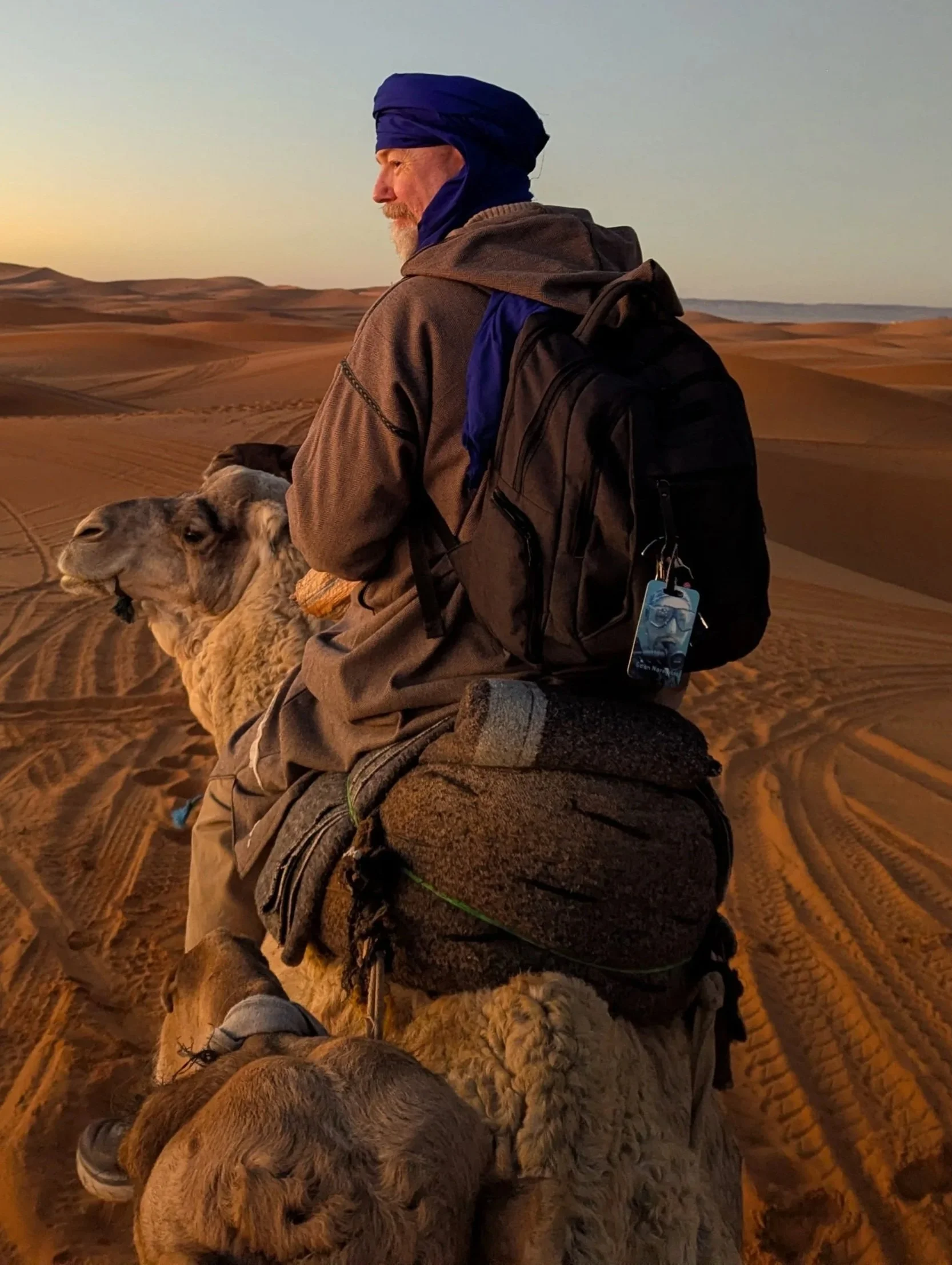 A man with a beard wearing a blue headwrap and a brown jacket, sitting on a camel in a desert at sunset with sand dunes in the background. The camel is lying down, and the man's backpack is visible.