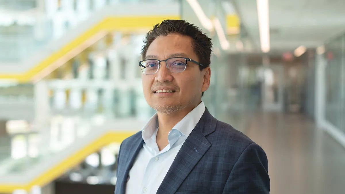 A professionally dressed man with glasses standing in a modern office building.