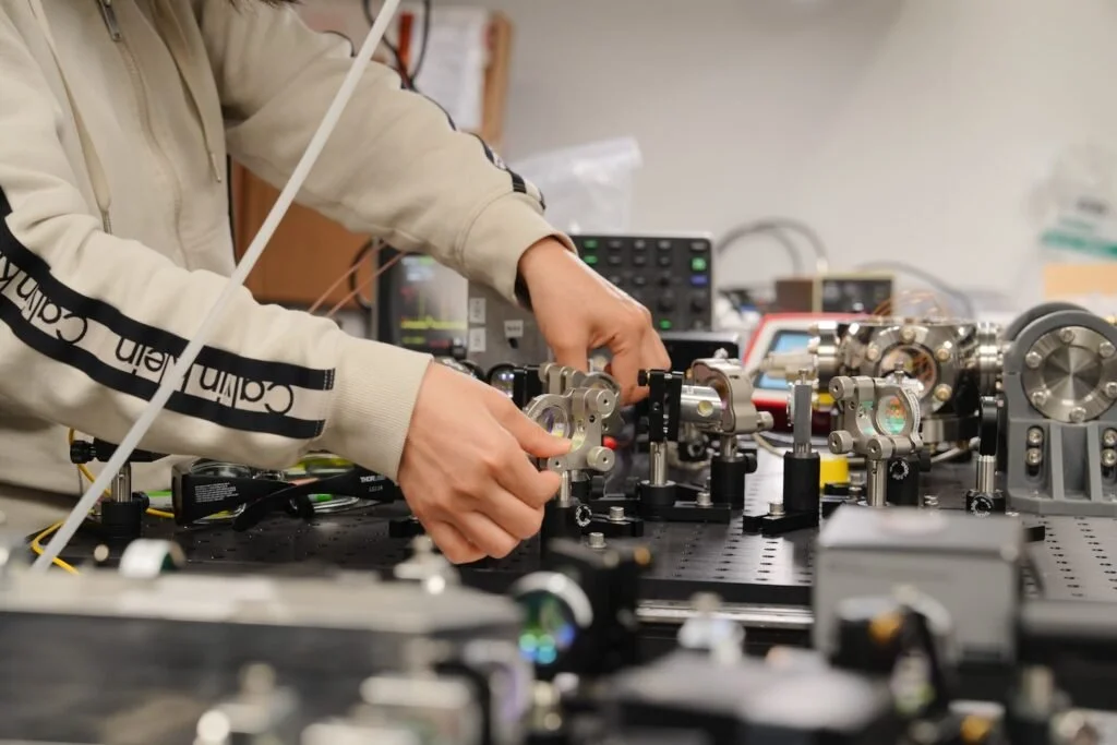 Person working on scientific equipment or experimental setup with metal components on a black perforated table.