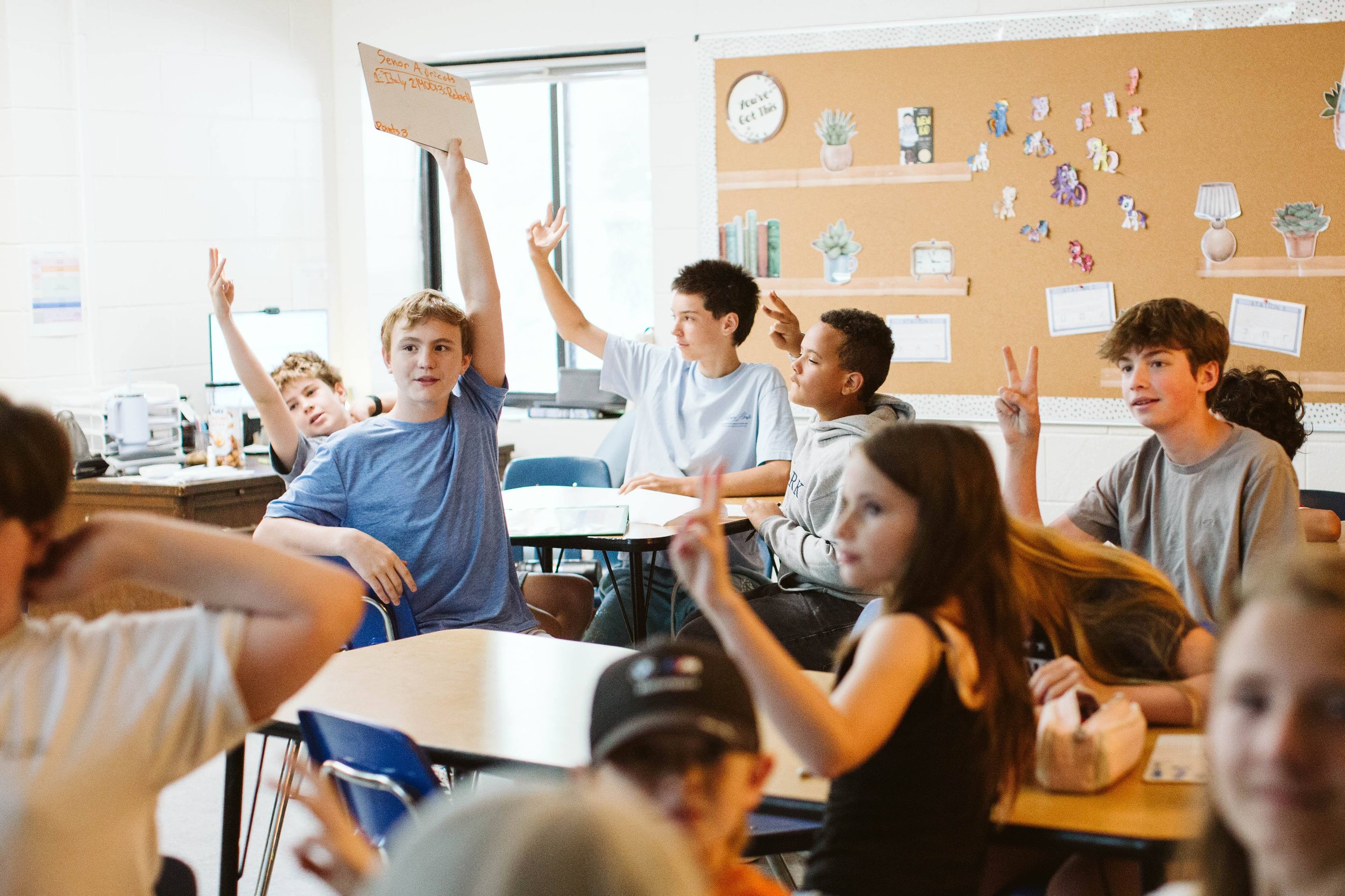 A classroom with multiple students seated at desks, some raising their hands. The students are diverse, and there is a bulletin board with decorations on the wall.