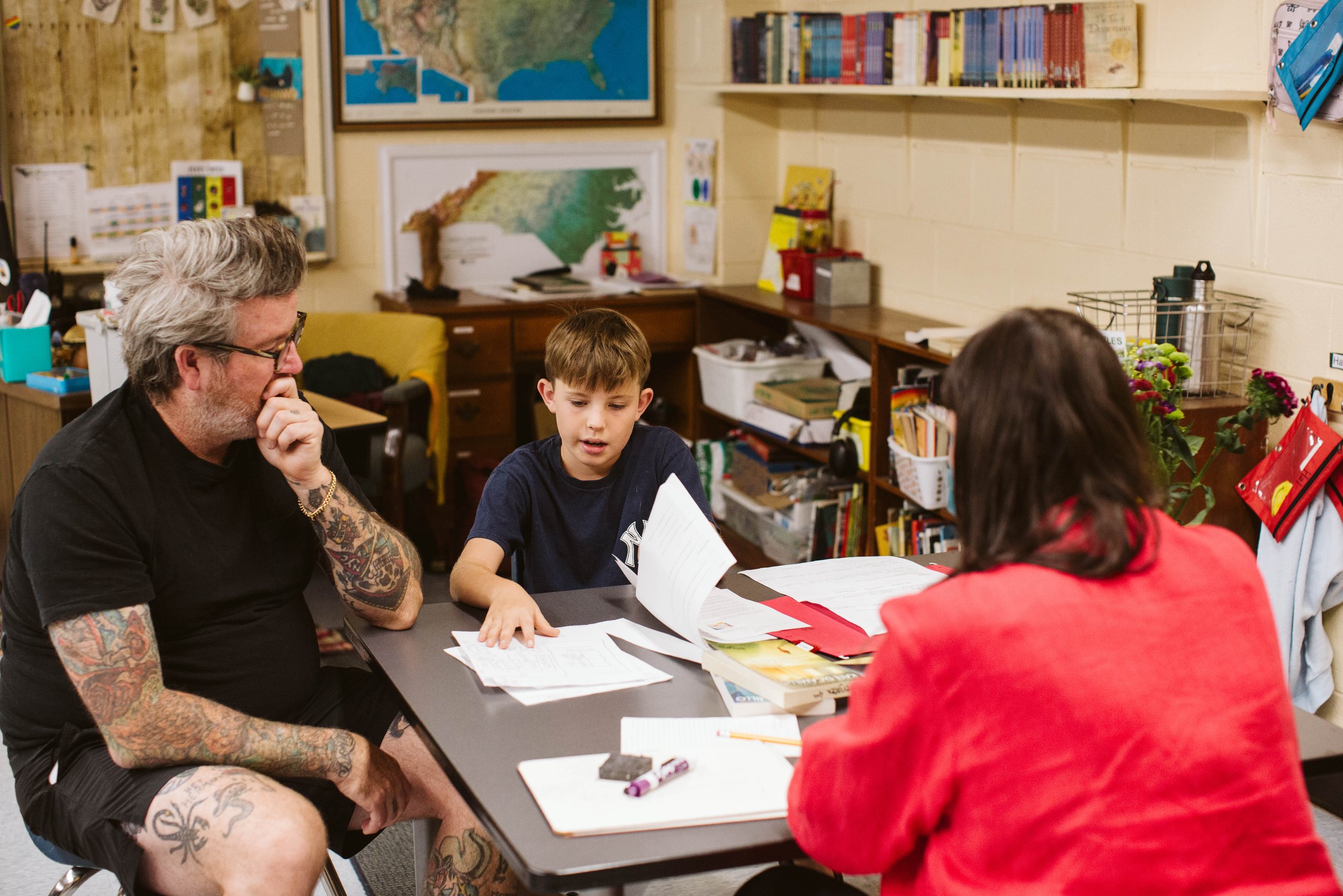 A man with tattoos and glasses sitting at a table with two children, a boy and a girl, in a classroom. The boy is reading a paper and the girl is seen from the back, wearing a red jacket. The room has educational posters, bookshelves, and a map of the United States on the wall.
