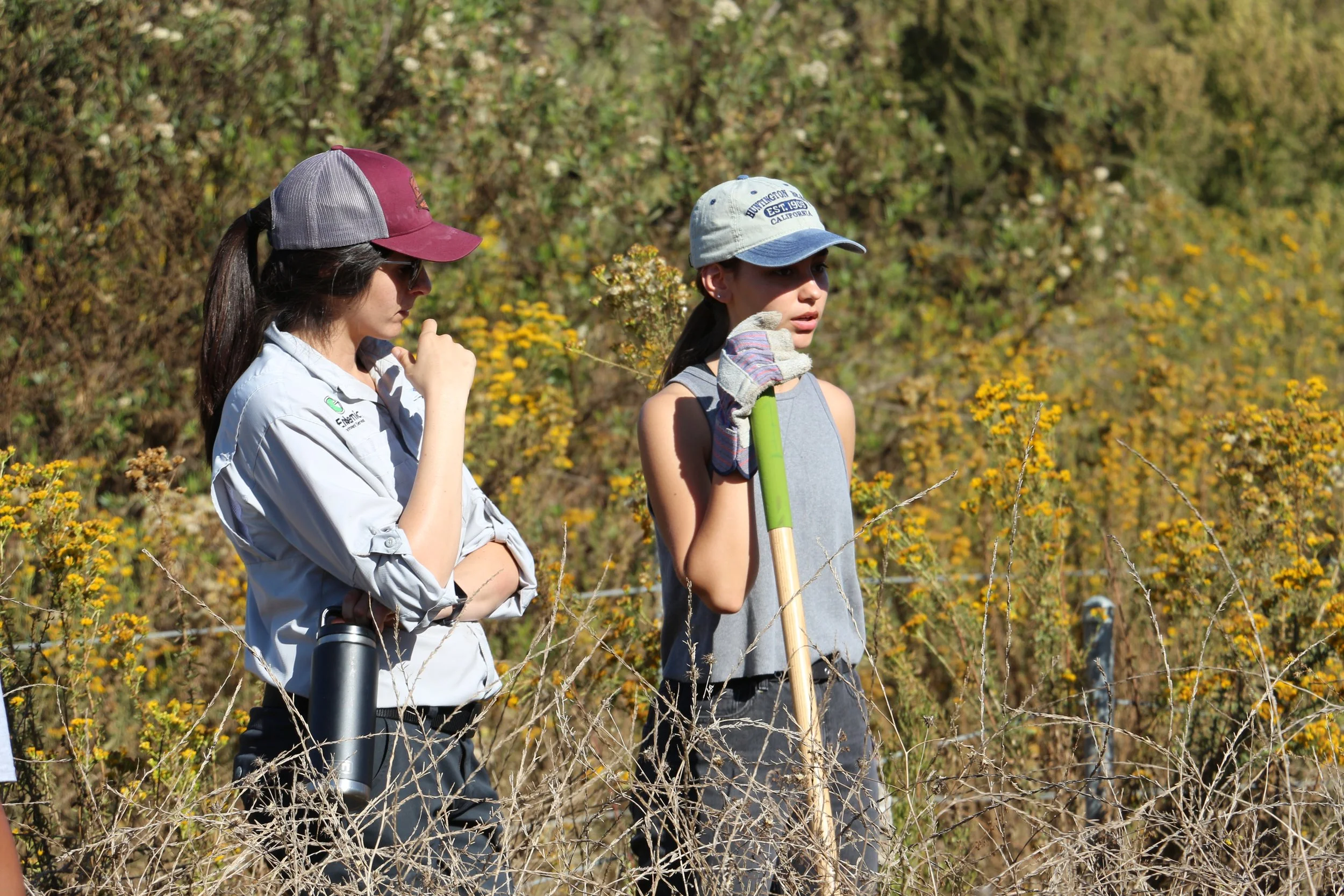 Two young women standing outdoors among yellow flowers and bushes, one wearing a gray and white cap and the other wearing a maroon and gray cap, both dressed in casual outdoor clothing, and holding gardening tools.