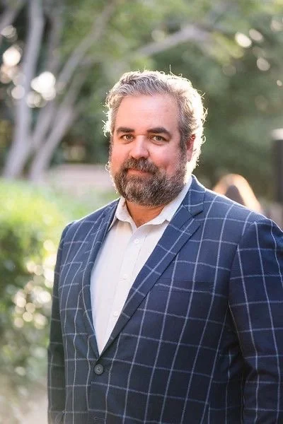 A man with gray hair and a beard in a blue checkered blazer stands outdoors with trees and grass in the background.