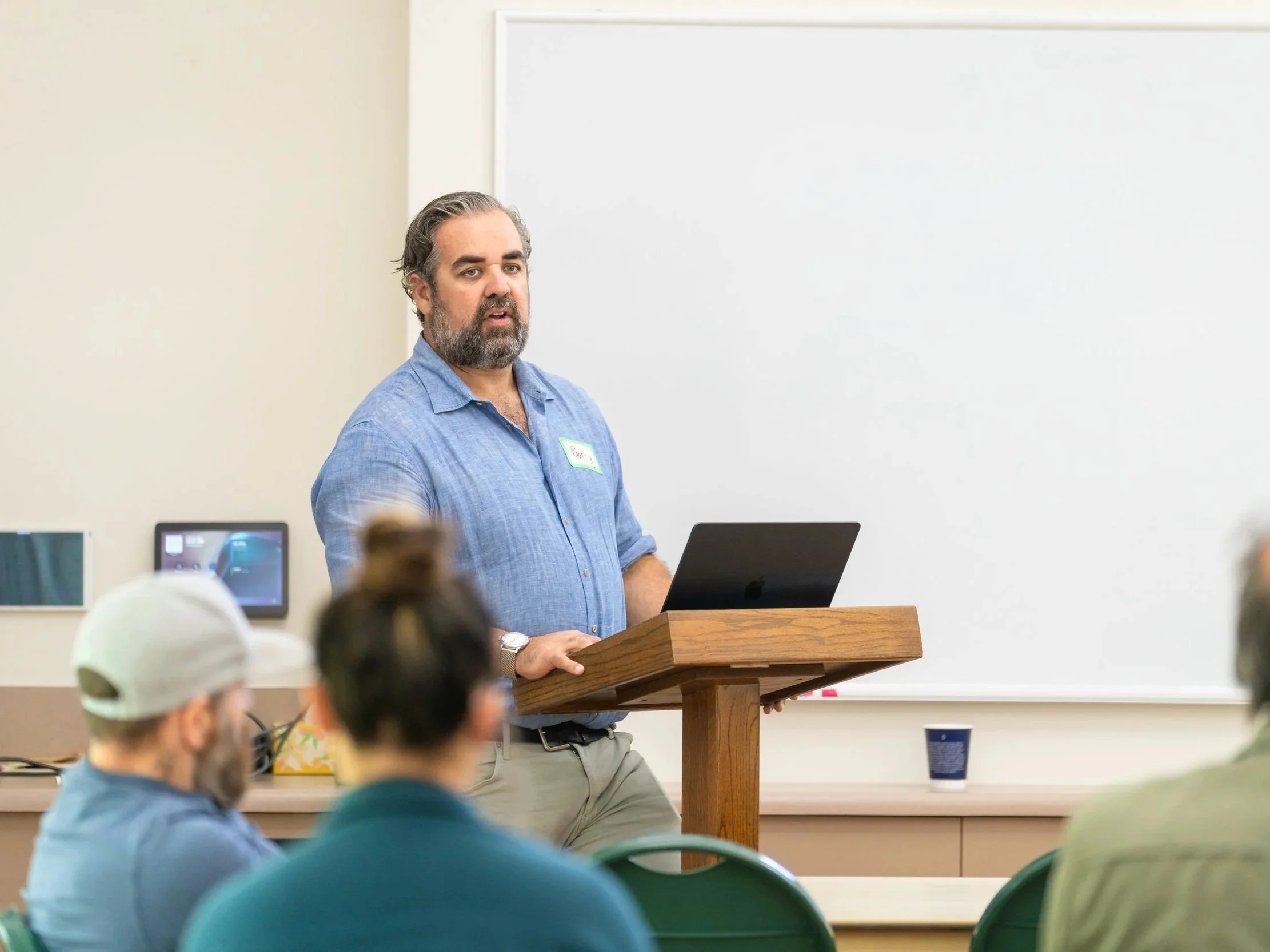 Man with gray hair and beard wearing a blue shirt giving a presentation in a classroom with students seated in front of him, some wearing hats.
