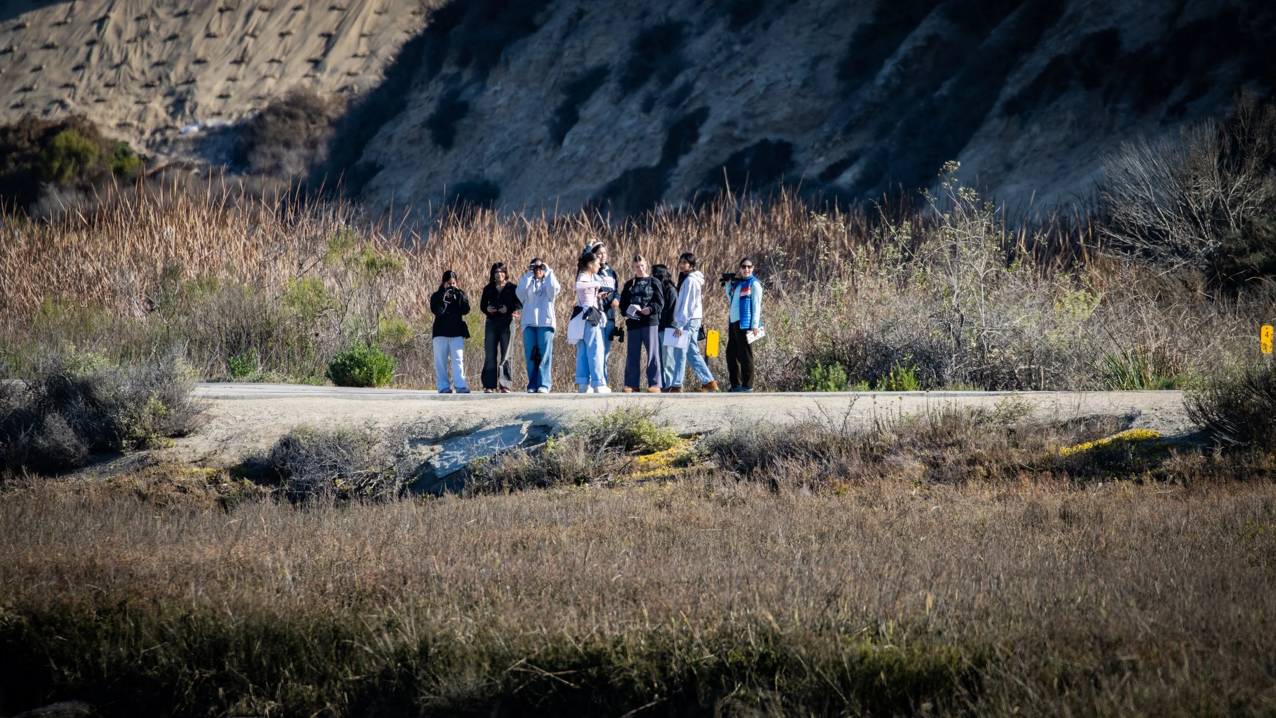 Group of people standing on the side of a paved road surrounded by dry grass and shrubs, with hills or mountains in the background.