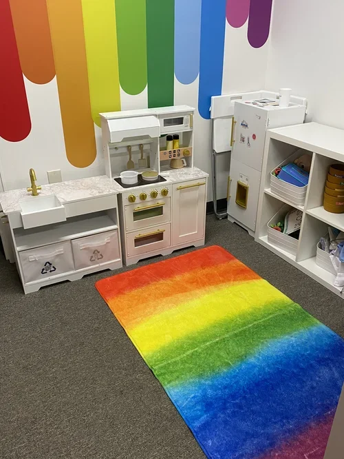 Colorful rainbow rug on gray carpet in a children's play kitchen area with white shelves and walls decorated with rainbow stripes.