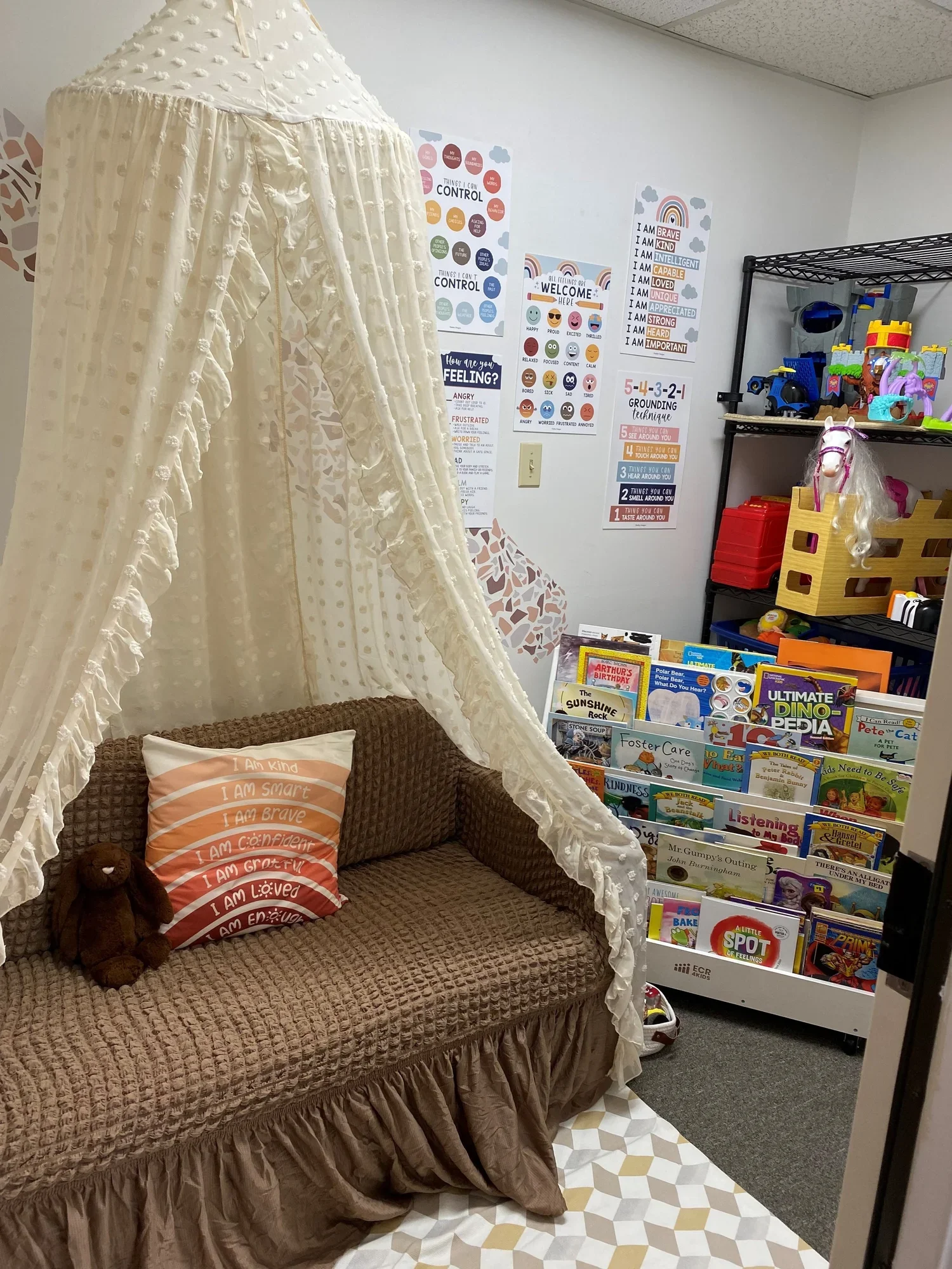 A cozy reading nook with a brown woven sofa, a ruffled bed skirt, and a pillow with positive affirmations. A small stuffed bear is on the sofa. There is a cream-colored canopy or tent with ruffled edges next to the sofa. The wall behind has various educational and motivational posters, and a bookshelf filled with children's books and toys. The floor has a geometric-patterned rug, and part of an adjacent room with carpet and shelves is visible.