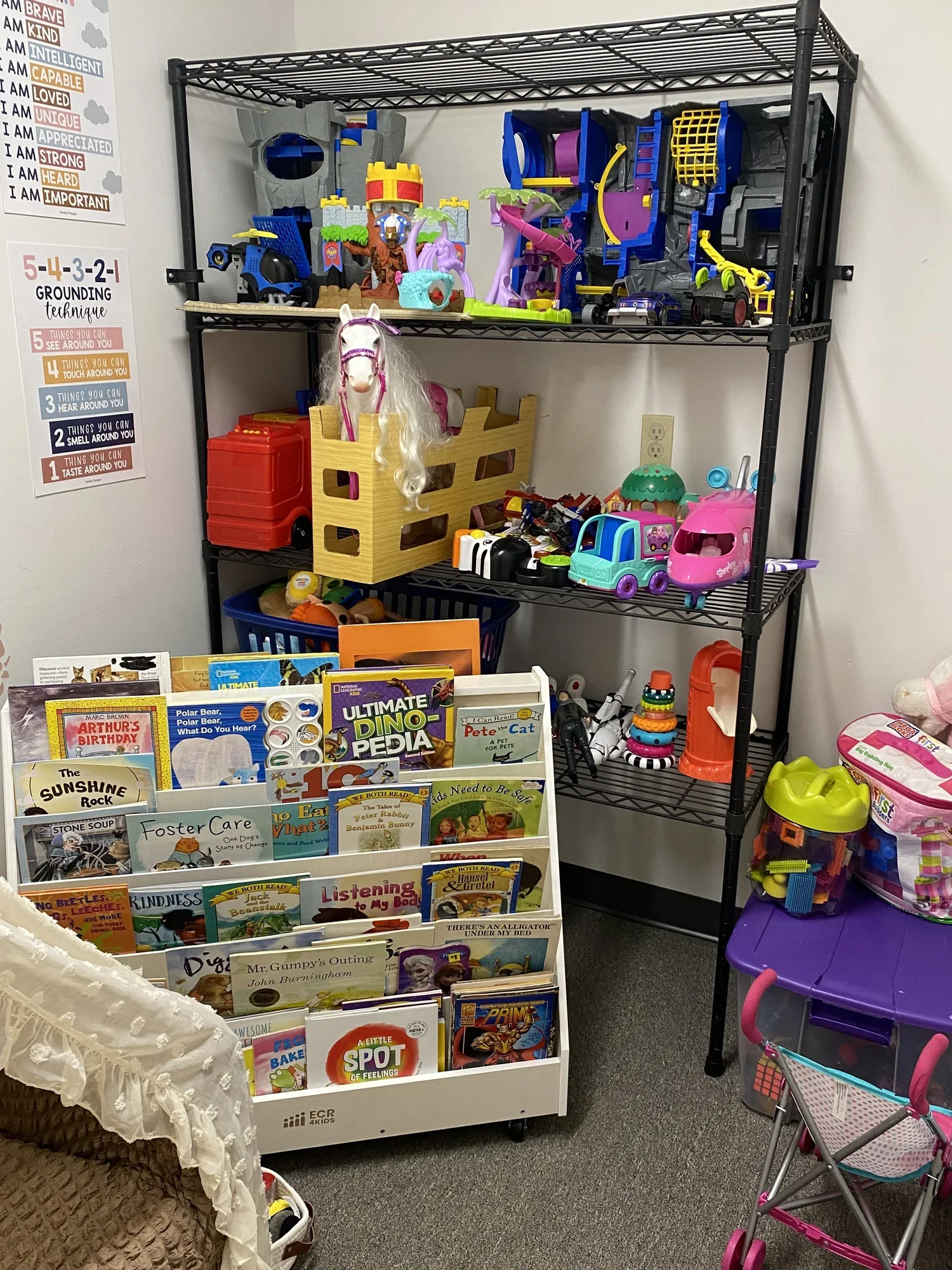 A display of children's toys and books on a black metal shelving unit and a white bookstand. The toys include plastic vehicles, a toy horse, and playsets. The books are children's picture books about animals and stories. There is a small pink stroller and colorful toys on the lower shelves.