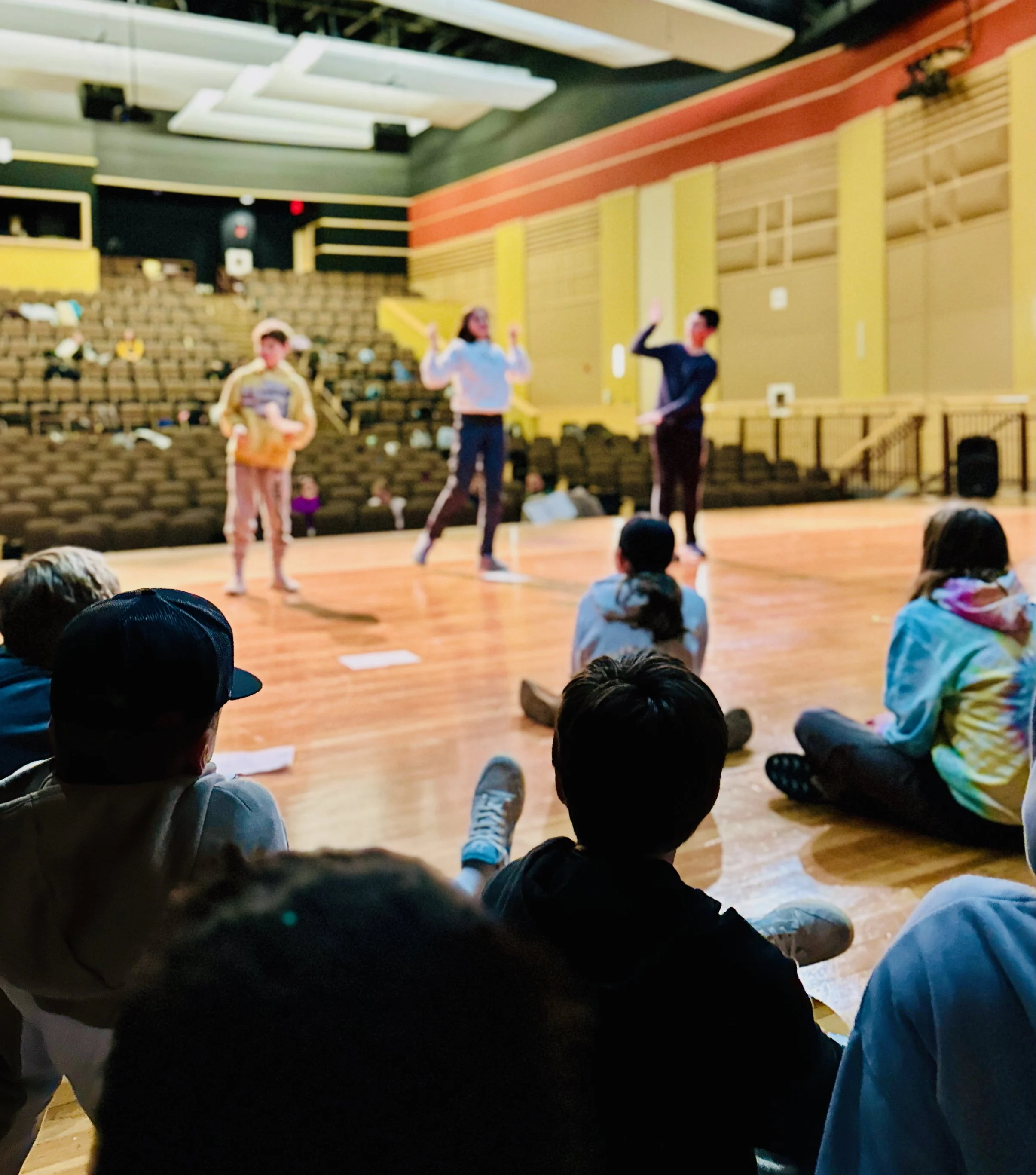 People in a theater or auditorium, some standing and some seated, with a focus on a group of three individuals on stage, engaged in a performance or presentation, with an audience visible in the foreground.