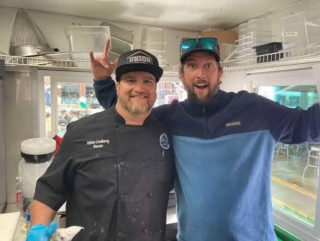 Dillon and Beau Lindborg, two men smiling inside a restaurant or food truck, one in a chef's uniform and the other in casual clothing, making rock and roll hand gestures.