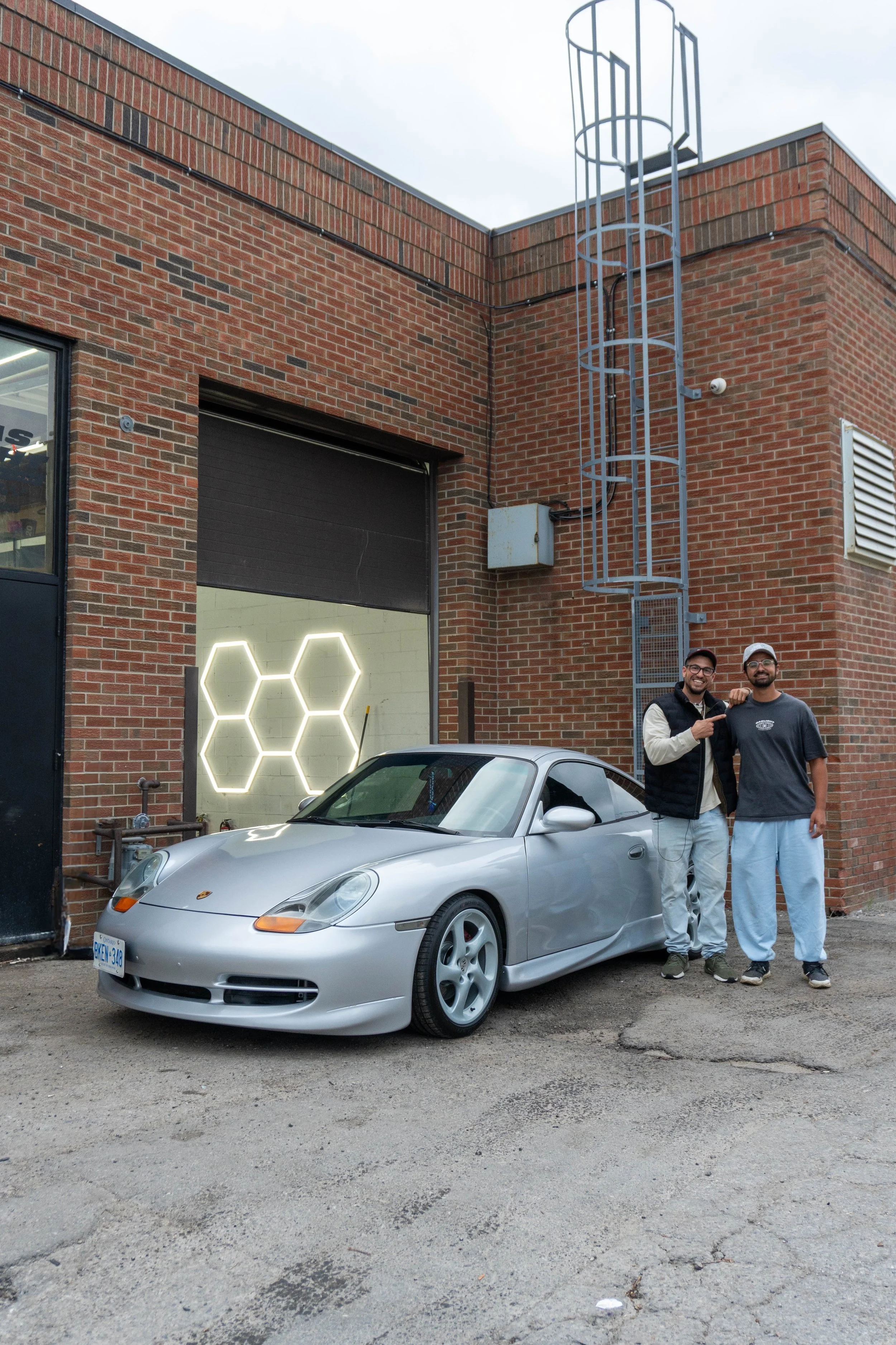Two young men standing next to a silver sports car parked outside a brick building with a closed garage door. The building has industrial elements like a metal ladder, vent, and an electrical box. The men are smiling, with one making a fist and the other with his arm around his friend's shoulder.