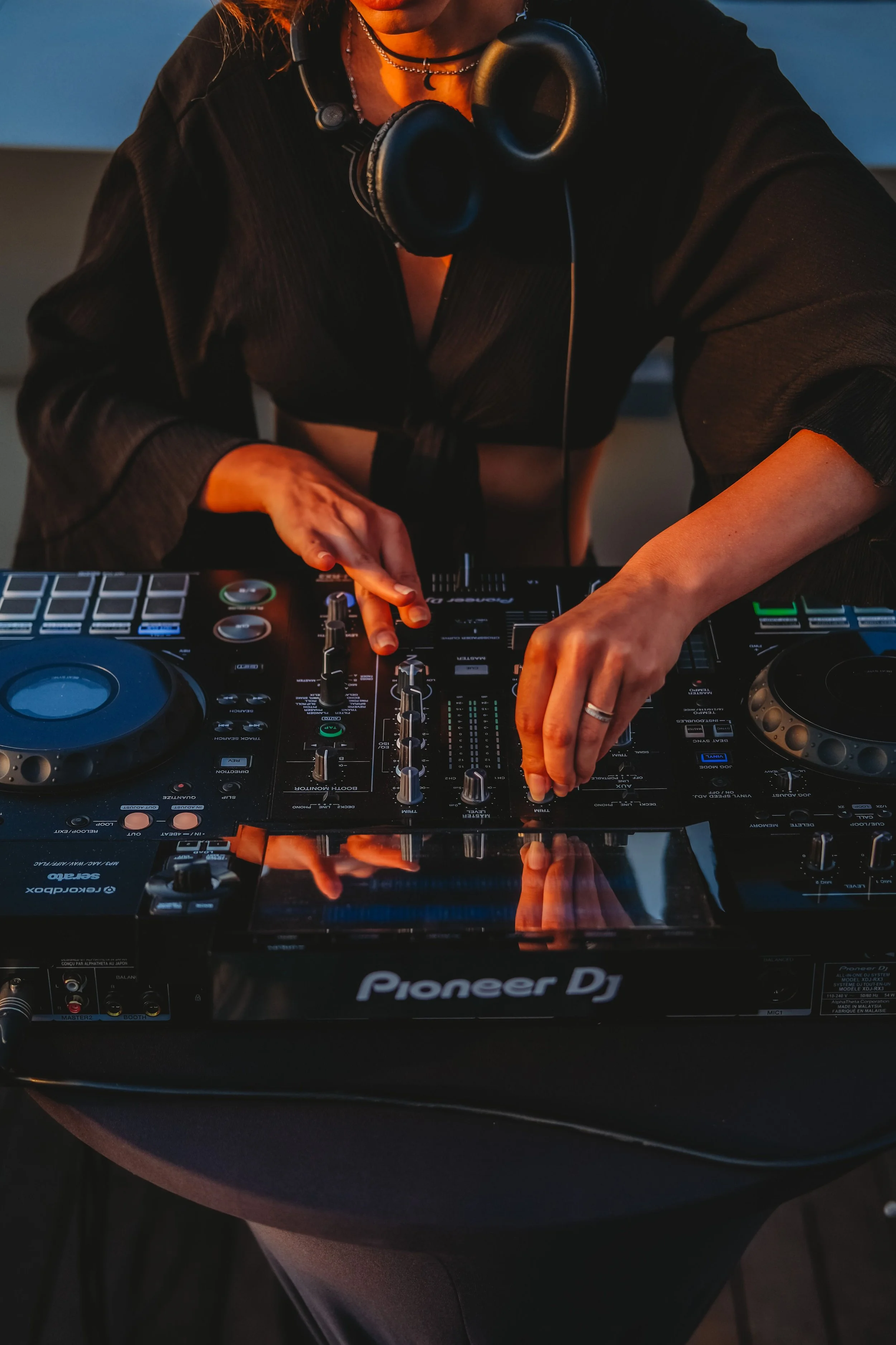 A DJ wearing a black shirt, with headphones around their neck, is operating a Pioneer DJ controller on a table. The scene is lit with warm, orange-toned lighting.