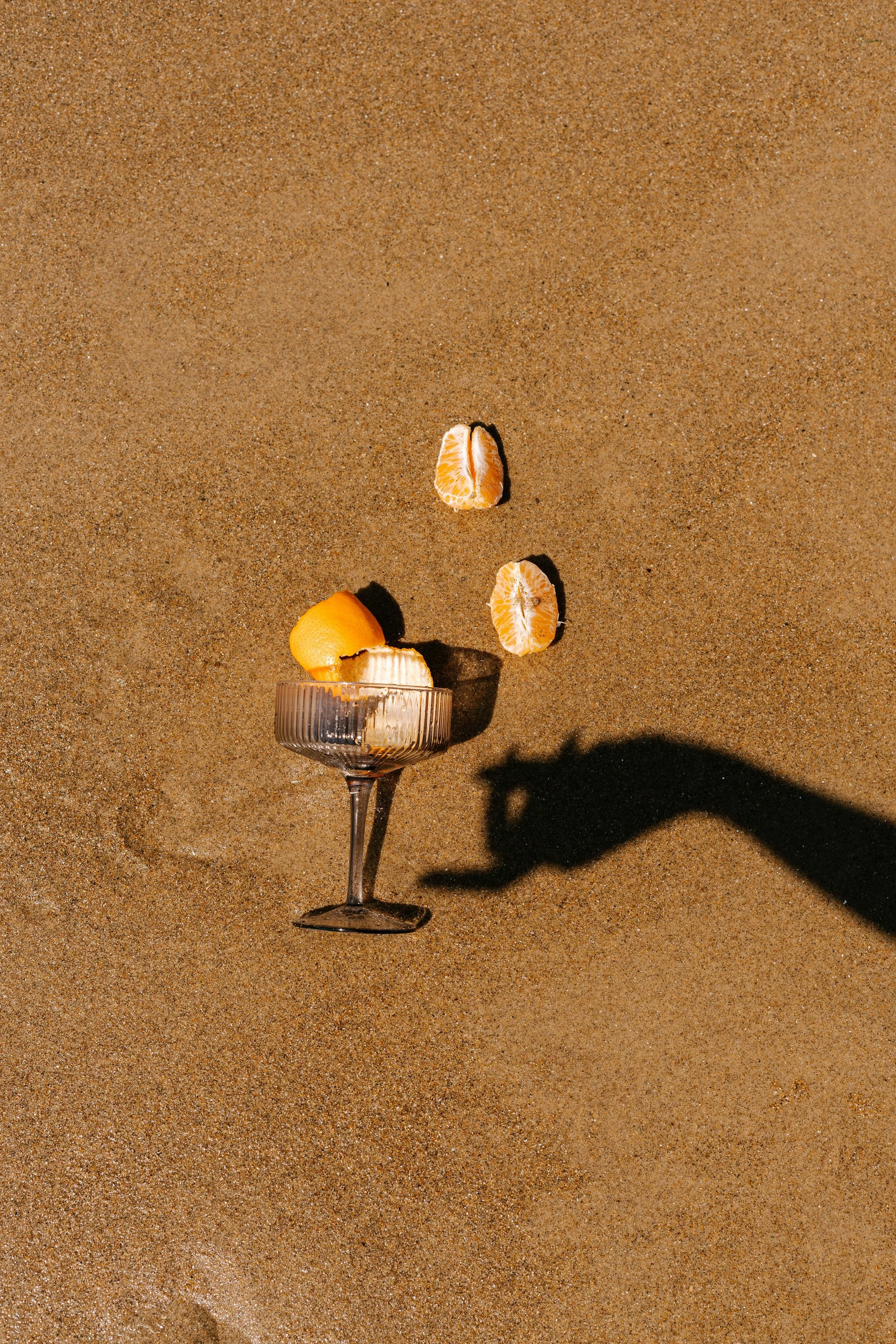 A clear wine glass filled with a peeled orange and two peeled orange segments, placed on a sandy beach with a plain background.